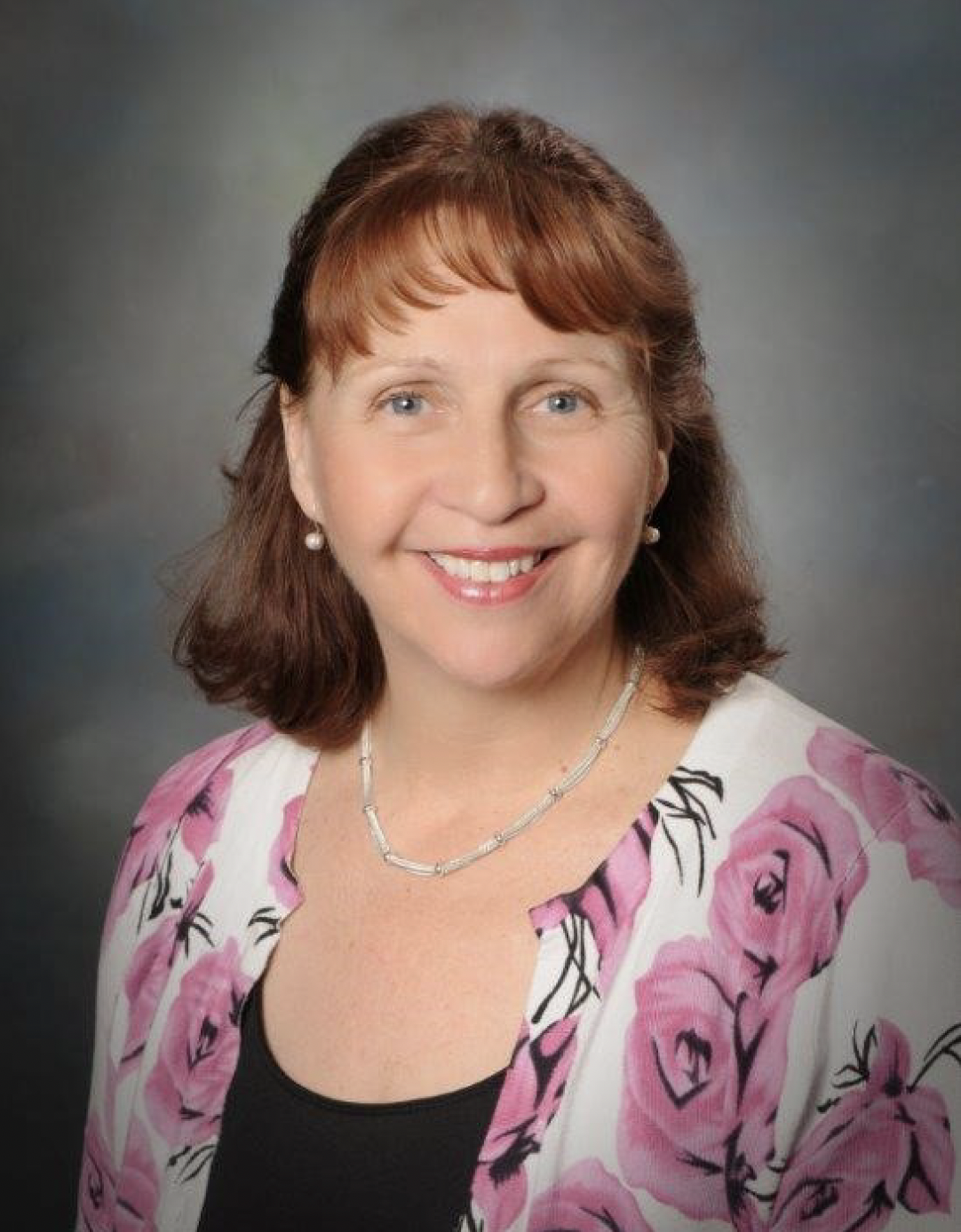 A middle-aged woman with shoulder-length brown hair and blue eyes smiling, wearing a floral white blazer with pink flowers, black top, pearl earrings, and a silver necklace.
