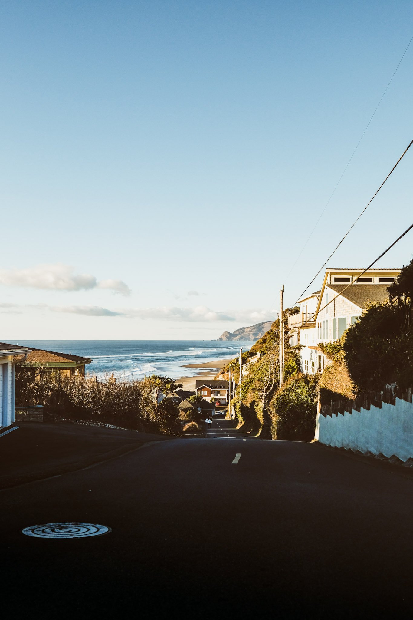A street leading down towards the beach with houses on the sides, ocean waves on the horizon, and a cliff in the distance under a blue sky.