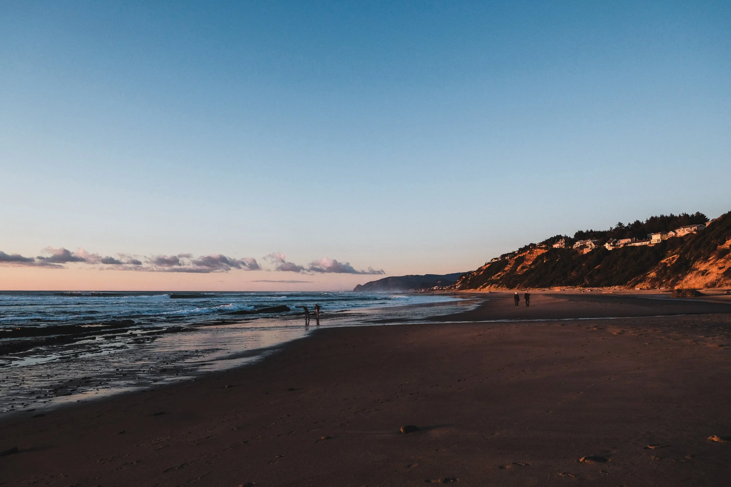 A beach at sunset with a few people walking along the shoreline, cliffs with houses on top in the background, and calm ocean waves.