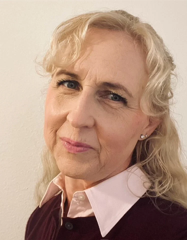 Close-up of a middle-aged woman with blonde curly hair, wearing a light pink collared shirt and a dark sweater, standing against a plain wall.