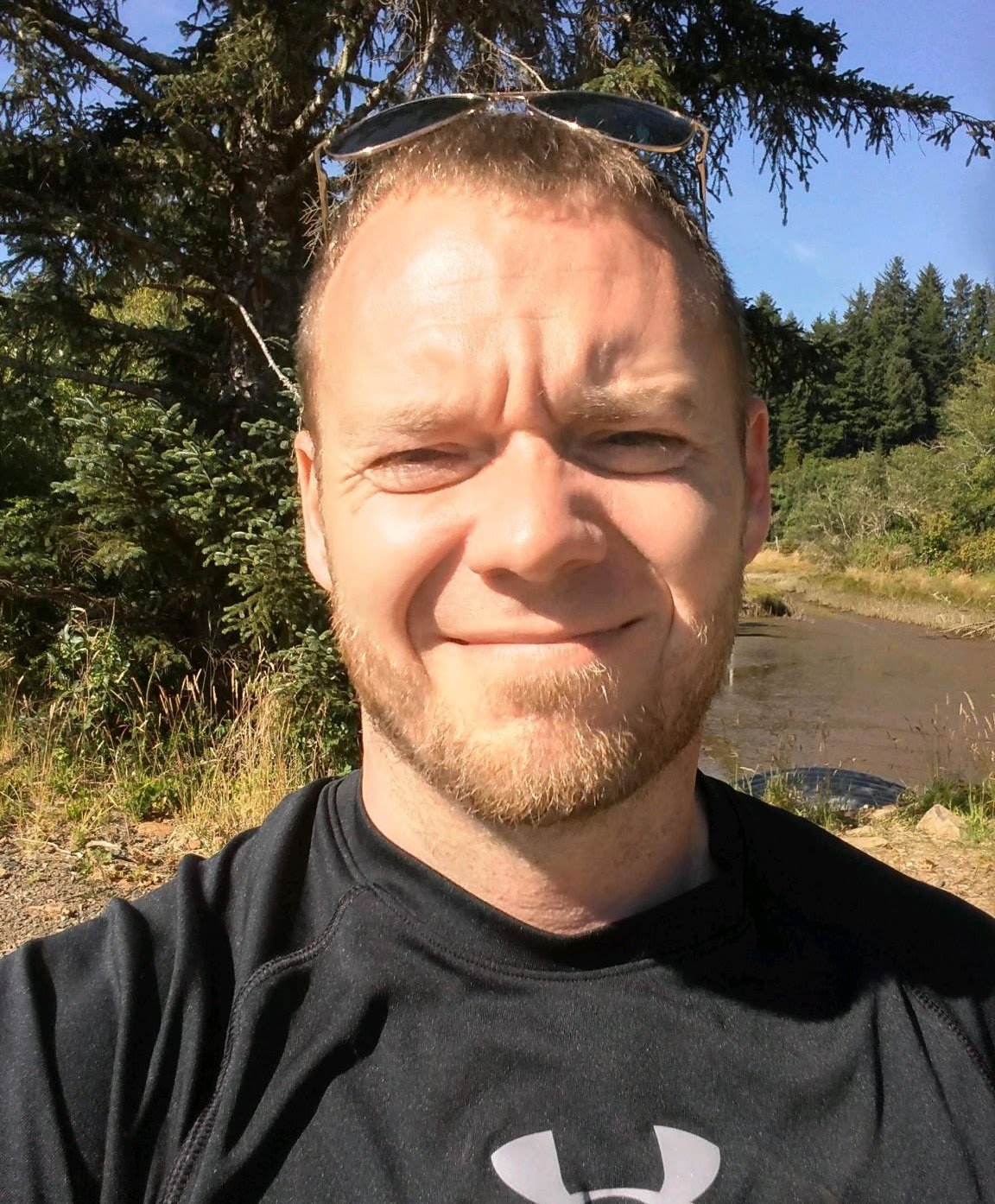 A man with short hair, beard, and sunglasses on his head taking a selfie outdoors with trees, bushes, and a river in the background.