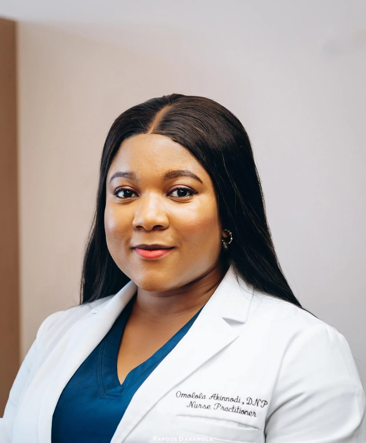 Headshot of a woman in medical attire, wearing a white coat with embroidery that reads 'Omolola Akinnobi, DNP, Nurse Practitioner', and a dark blue top underneath. She has long black hair, styled straight, and is smiling softly, standing against a plain background.