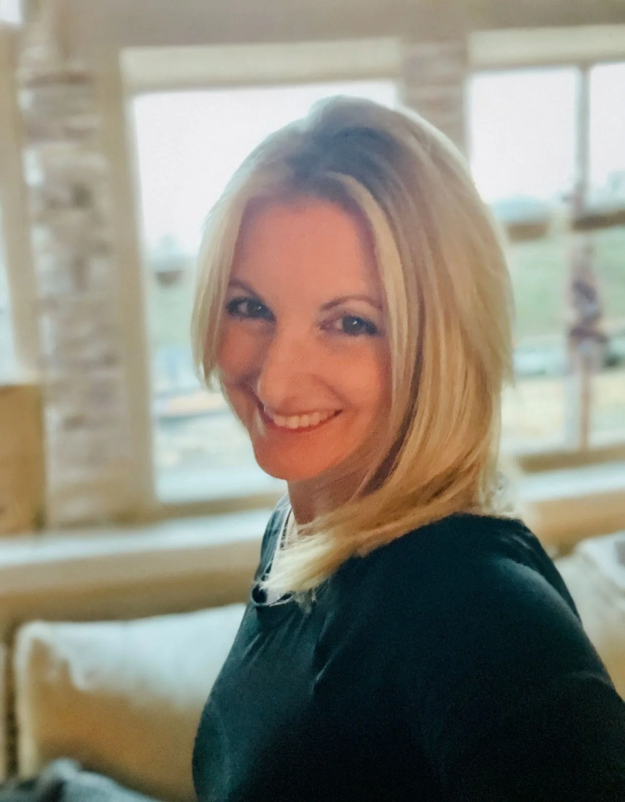 A woman with blonde hair smiling inside a room with large windows and natural light.