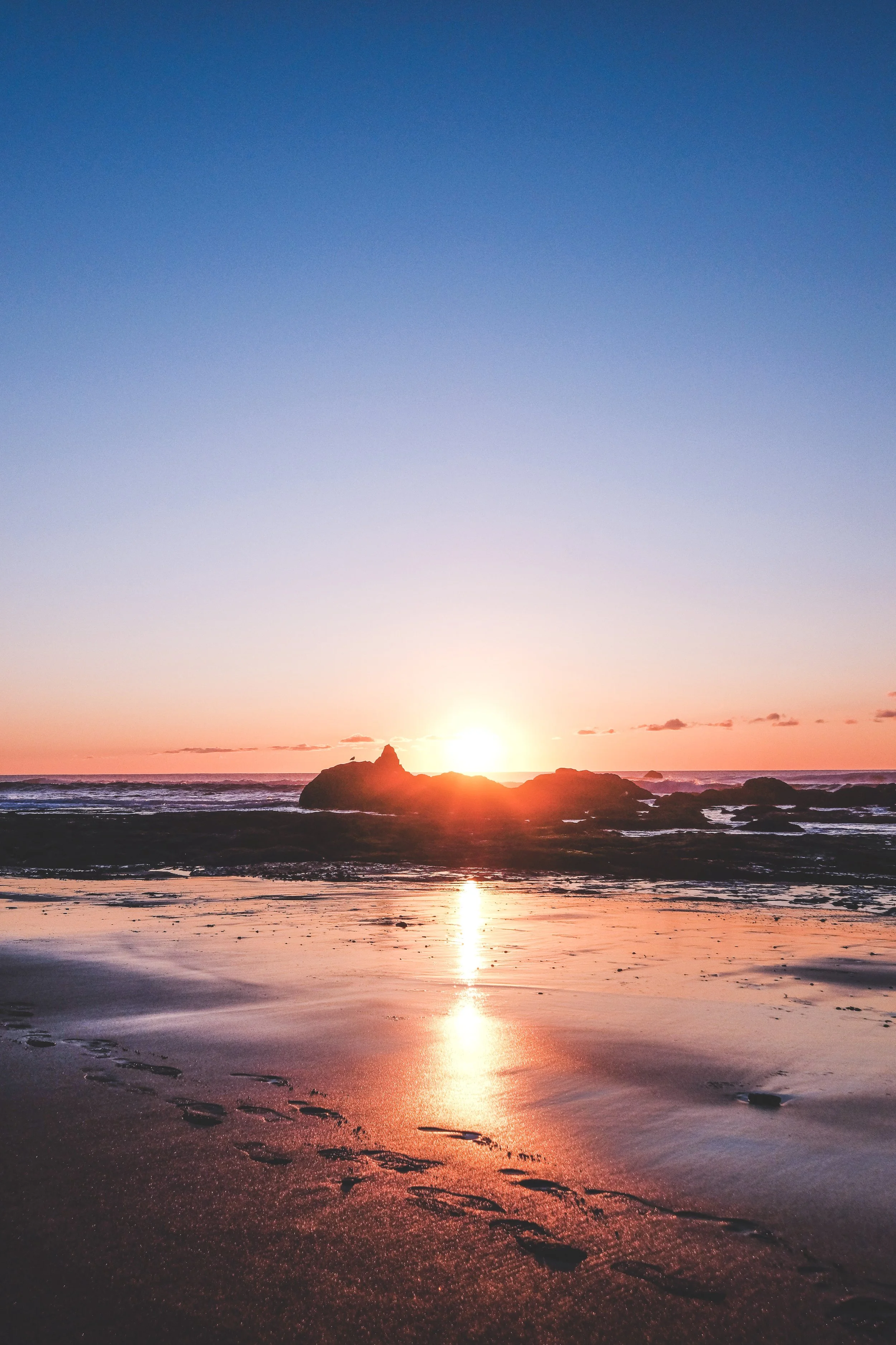 Sunset over a rocky beach, with the sun partially behind rocks and its reflection on the wet sand.