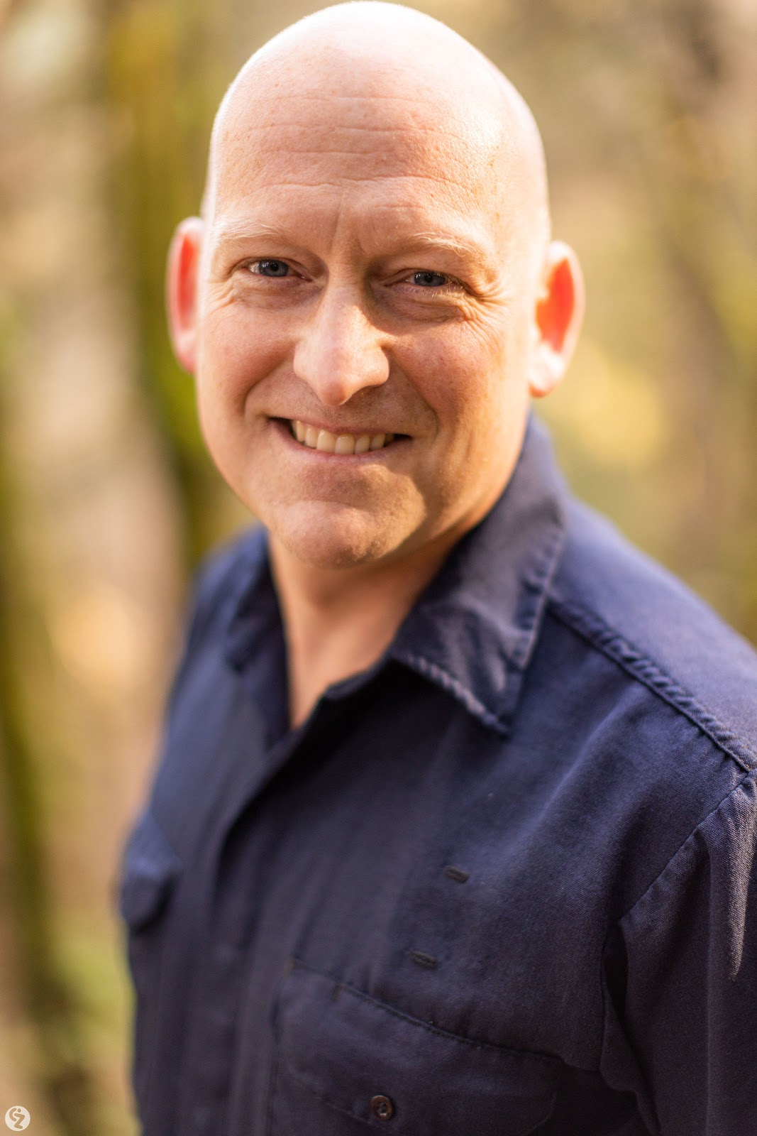 A smiling man with a bald head, light skin, and blue eyes, wearing a dark navy button-up shirt, standing outdoors with blurred trees and foliage in the background.