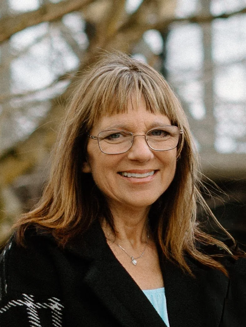 A woman with light brown hair, glasses, and a black coat standing outdoors near a tree, smiling at the camera.