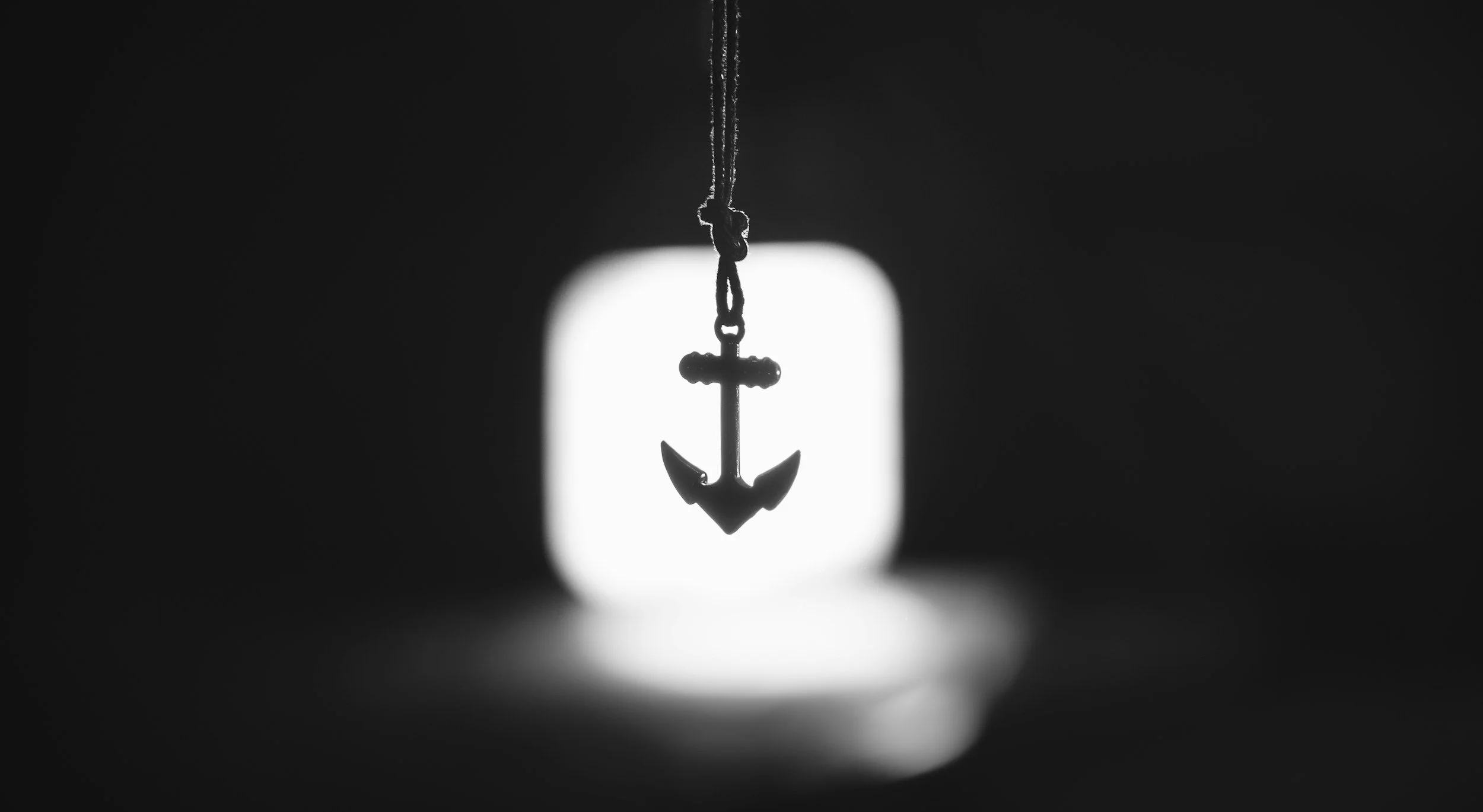 Silhouette of an anchor hanging from a rope in front of a blurred, bright background.
