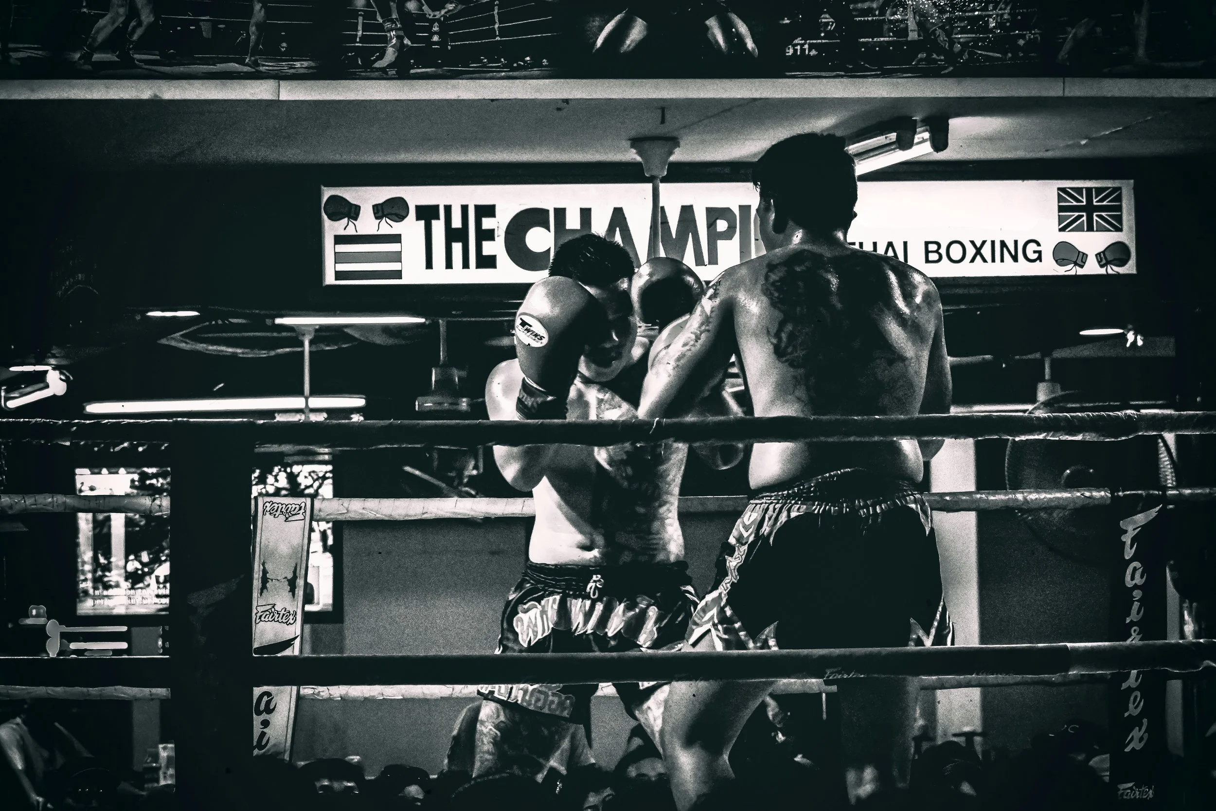 Two male boxers sparring in a boxing ring at a gym, with a sign that reads 'The Champion.'