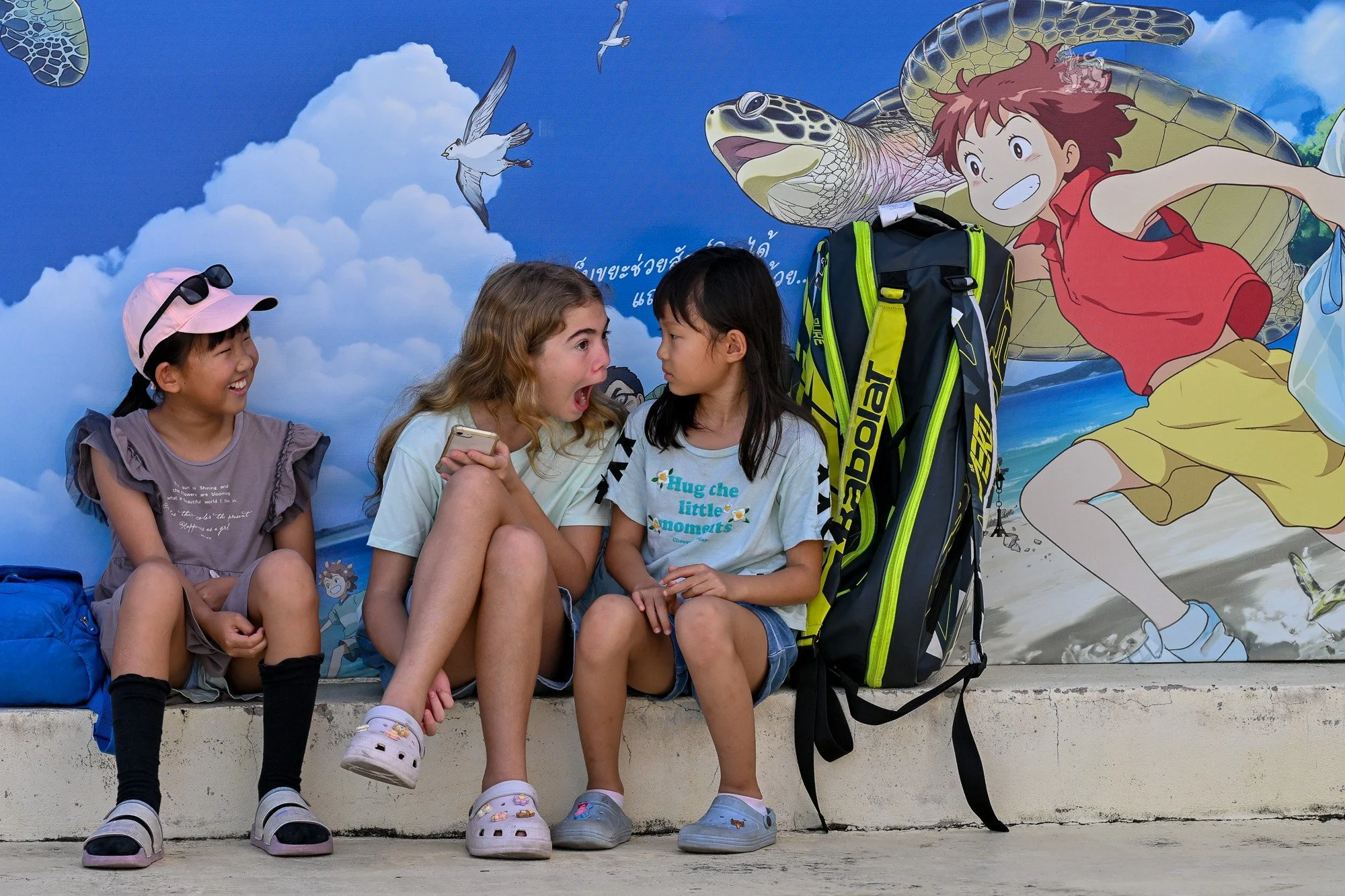 Three young girls sitting in front of a colorful mural, with one girl looking surprised or shouting, another girl smiling, and the third girl listening, as a large tennis bag is placed nearby.