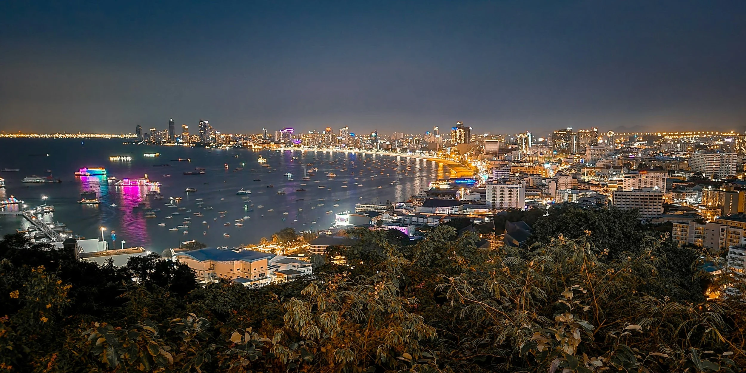 Nighttime cityscape of Bangkok with illuminated skyscrapers, boats on the Chao Phraya River, and lush greenery in the foreground.