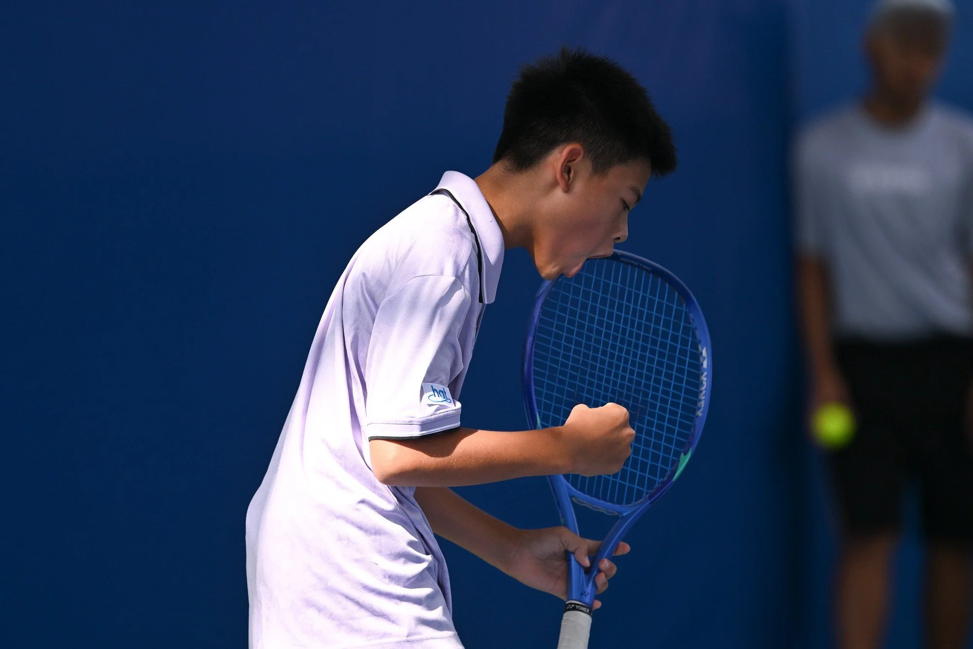 Young male tennis player in white uniform celebrating on the court, clutching a blue tennis racket, with another person holding a tennis ball in the background.