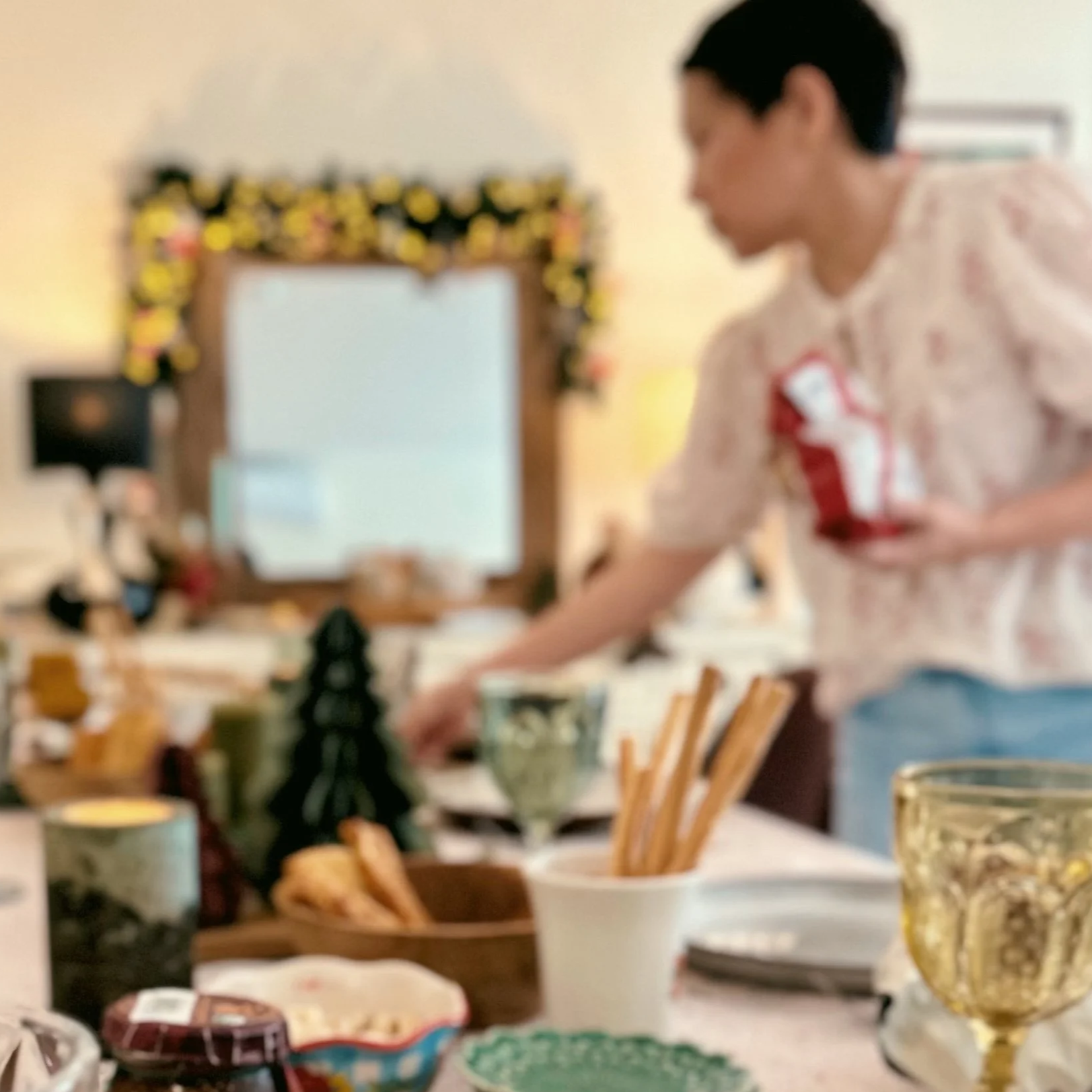 A woman with short dark hair wearing a white shirt with red print, standing in a dining or serving area. The background features a mirror decorated with a garland of flowers or lights, and various food items and dining accessories are visible on the table in the foreground.