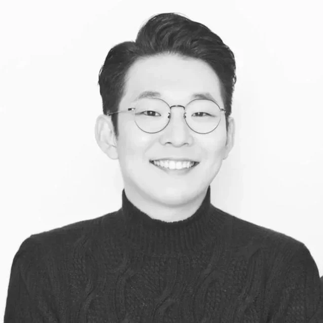 Portrait of a young man with short dark hair, wearing glasses, and a black turtleneck sweater, smiling in front of a plain background.