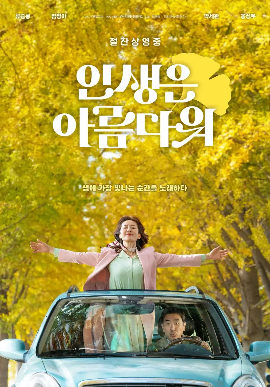 A woman with open arms stands through the sunroof of a car on a tree-lined road with yellow autumn leaves. A man is driving the car with a surprised expression.