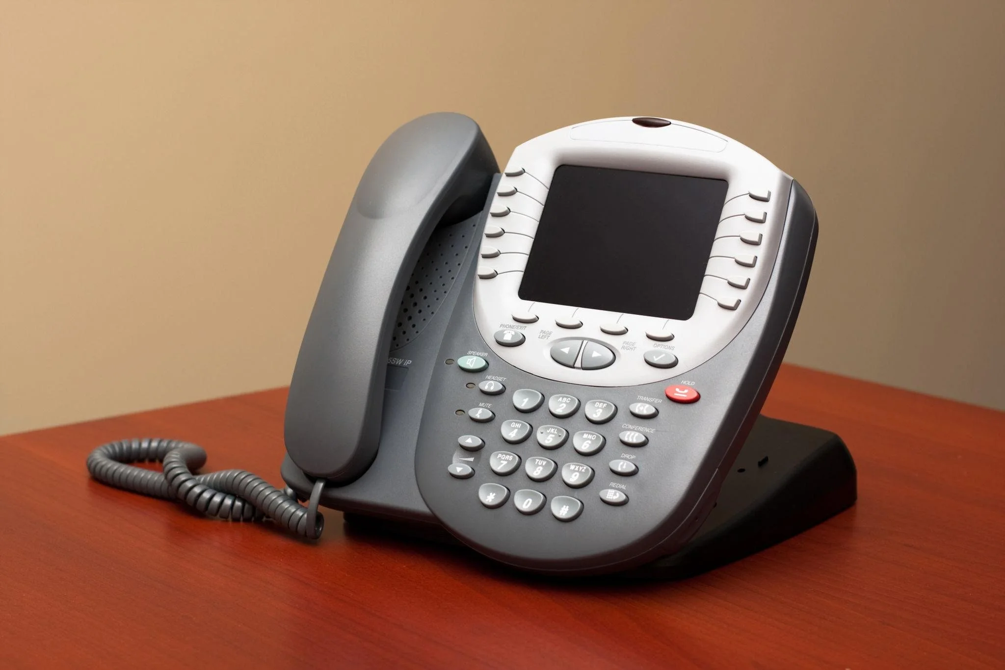 Office desk phone with cord, gray and white body, and keypad, placed on a wooden surface with a beige wall background.