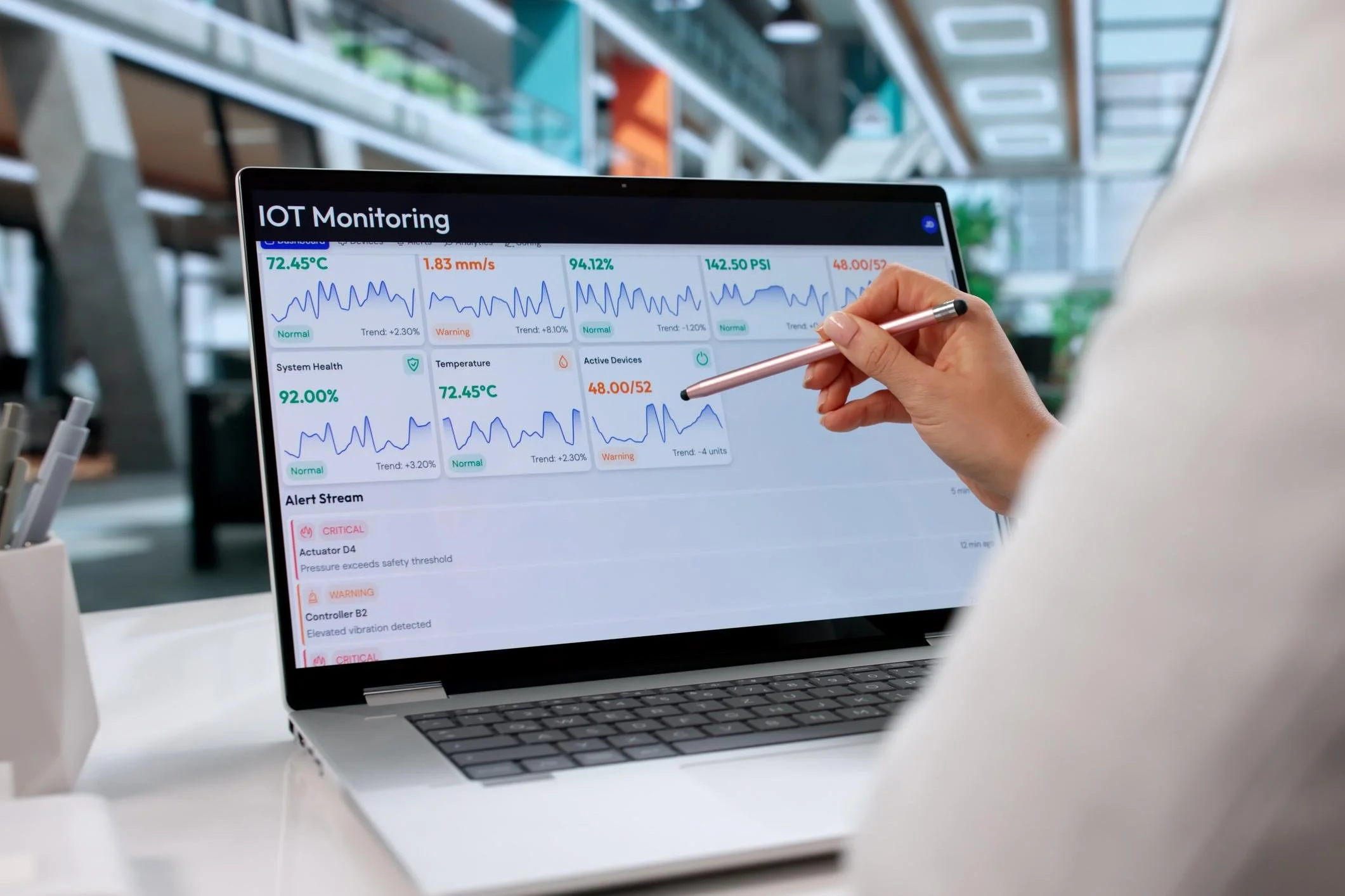 A person holding a stylus pointing at a laptop screen displaying an IoT monitoring dashboard with graphs, temperature, pressure, and system health indicators in a modern office environment.
