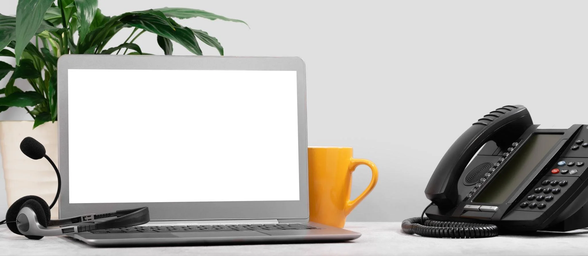 An office desk with a laptop, a headset, a yellow coffee mug, and a black landline phone. A green plant is in the background.