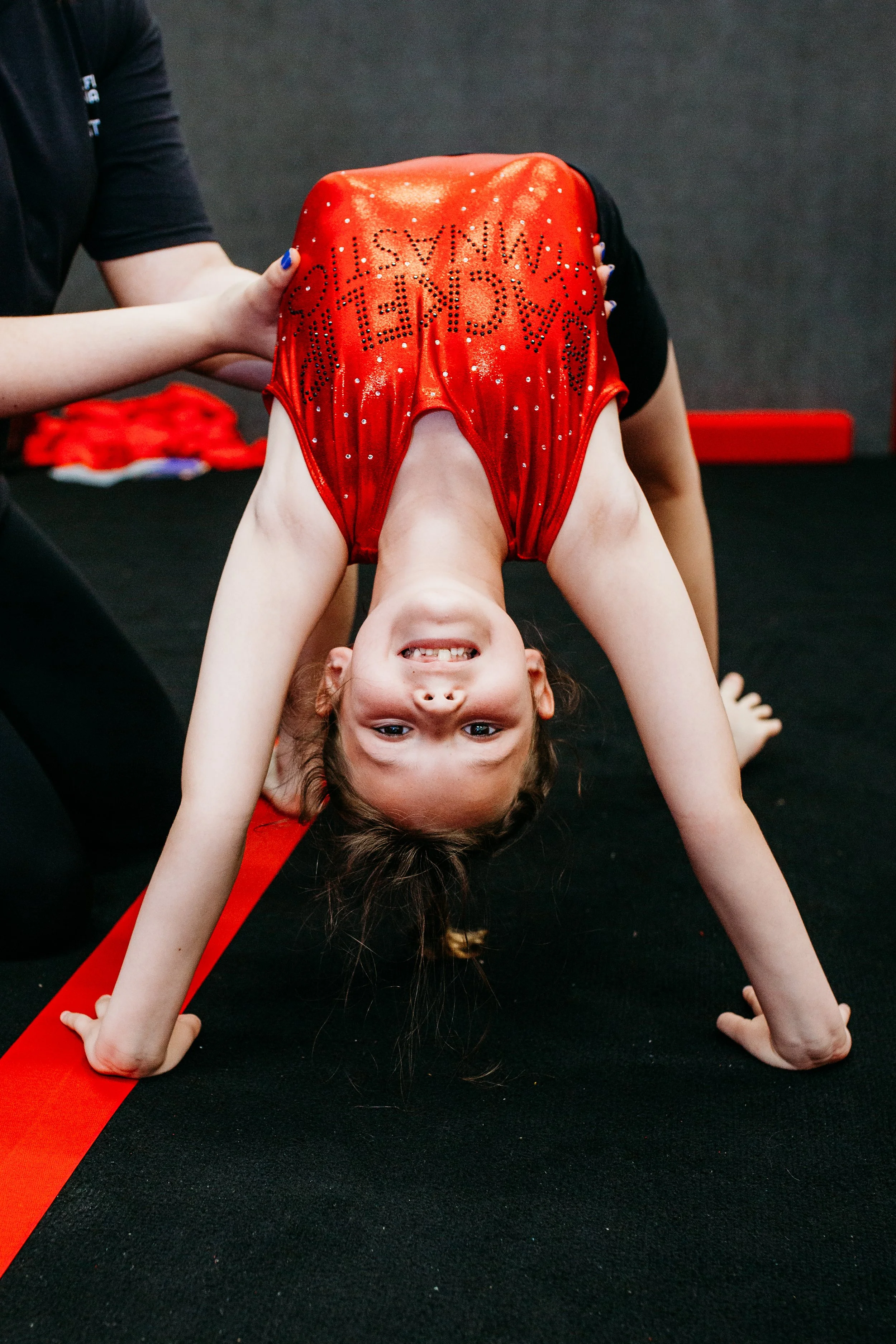 Young girl in a red gymnastics leotard doing a backbend on a black mat with red stripes, smiling and looking at the camera.