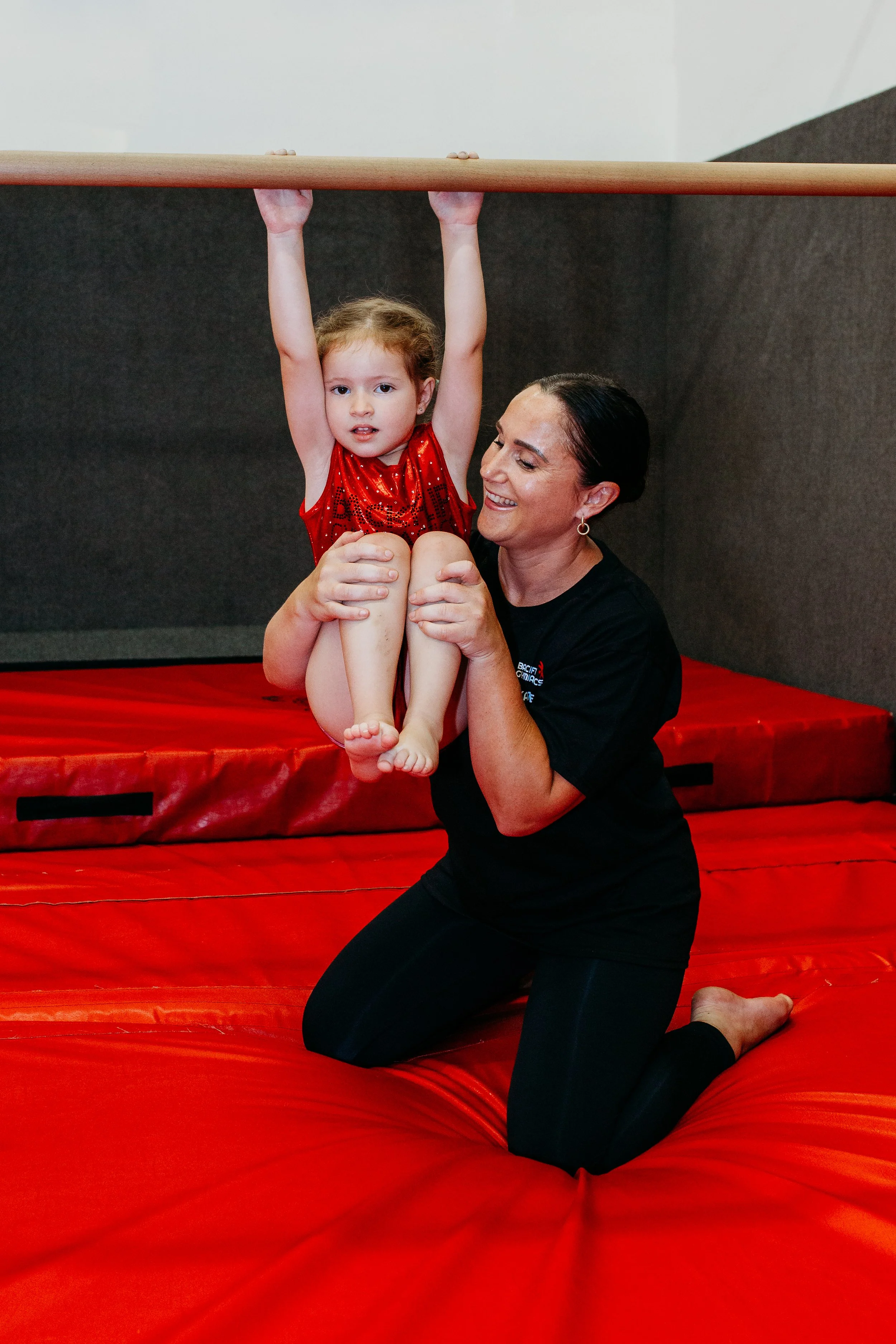 A young girl in a red dress hanging from a gymnastics bar with the assistance of a woman kneeling on a red mat.
