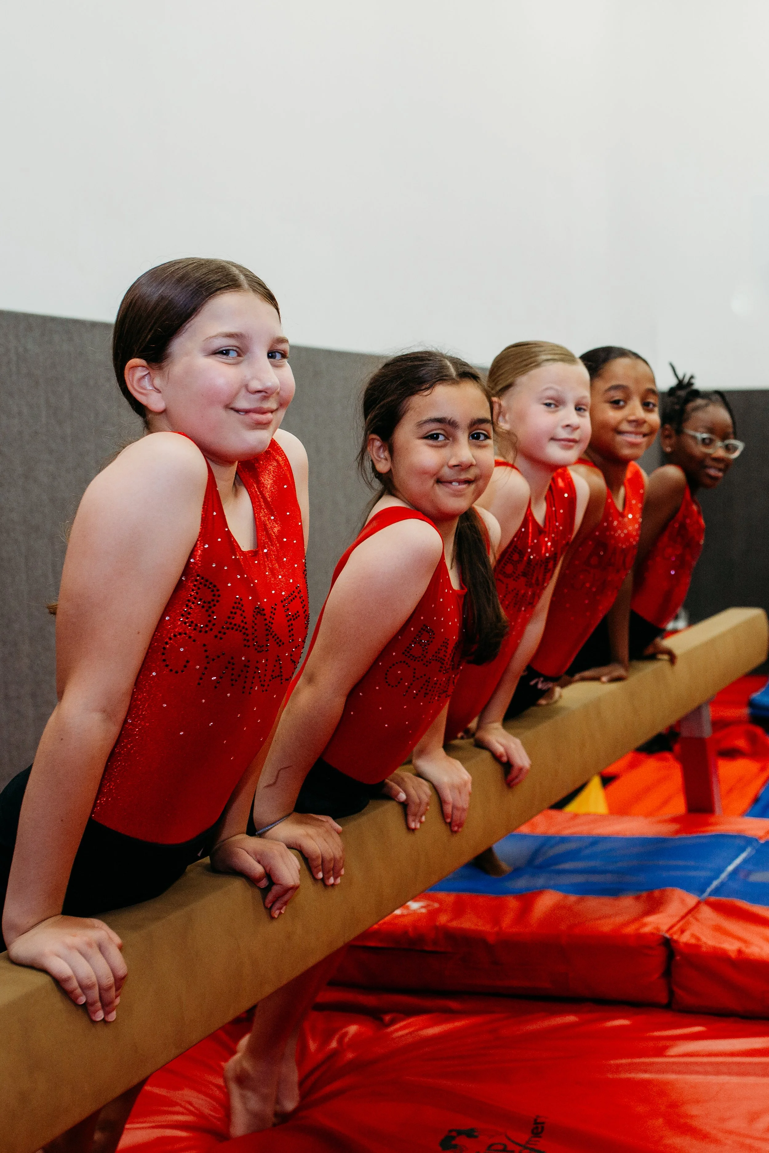 Five young girls in red gymnastics leotards practicing on a balance beam in a gymnastics facility.