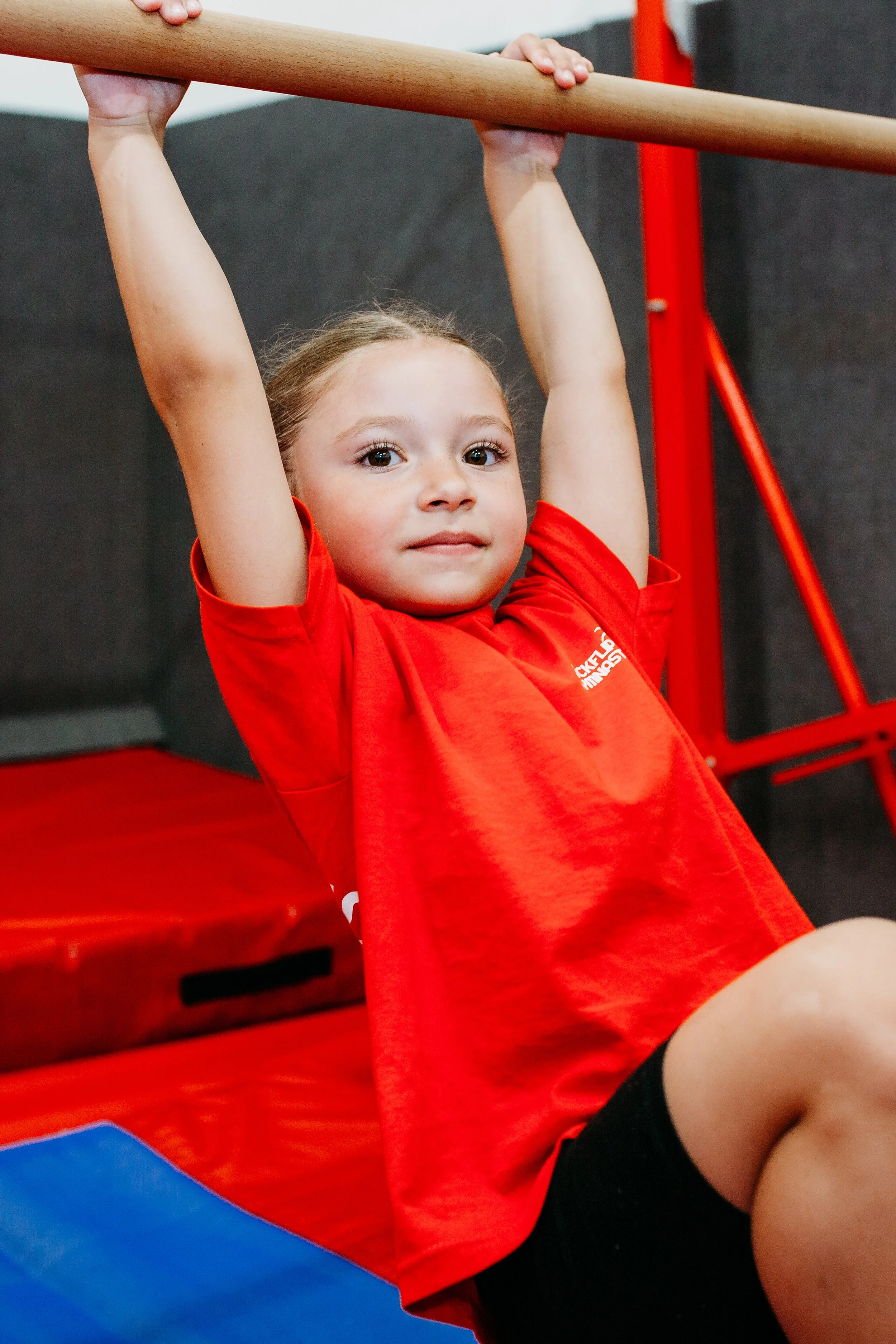 Young girl in a red t-shirt and black shorts hanging on a gymnastics pommel horse.