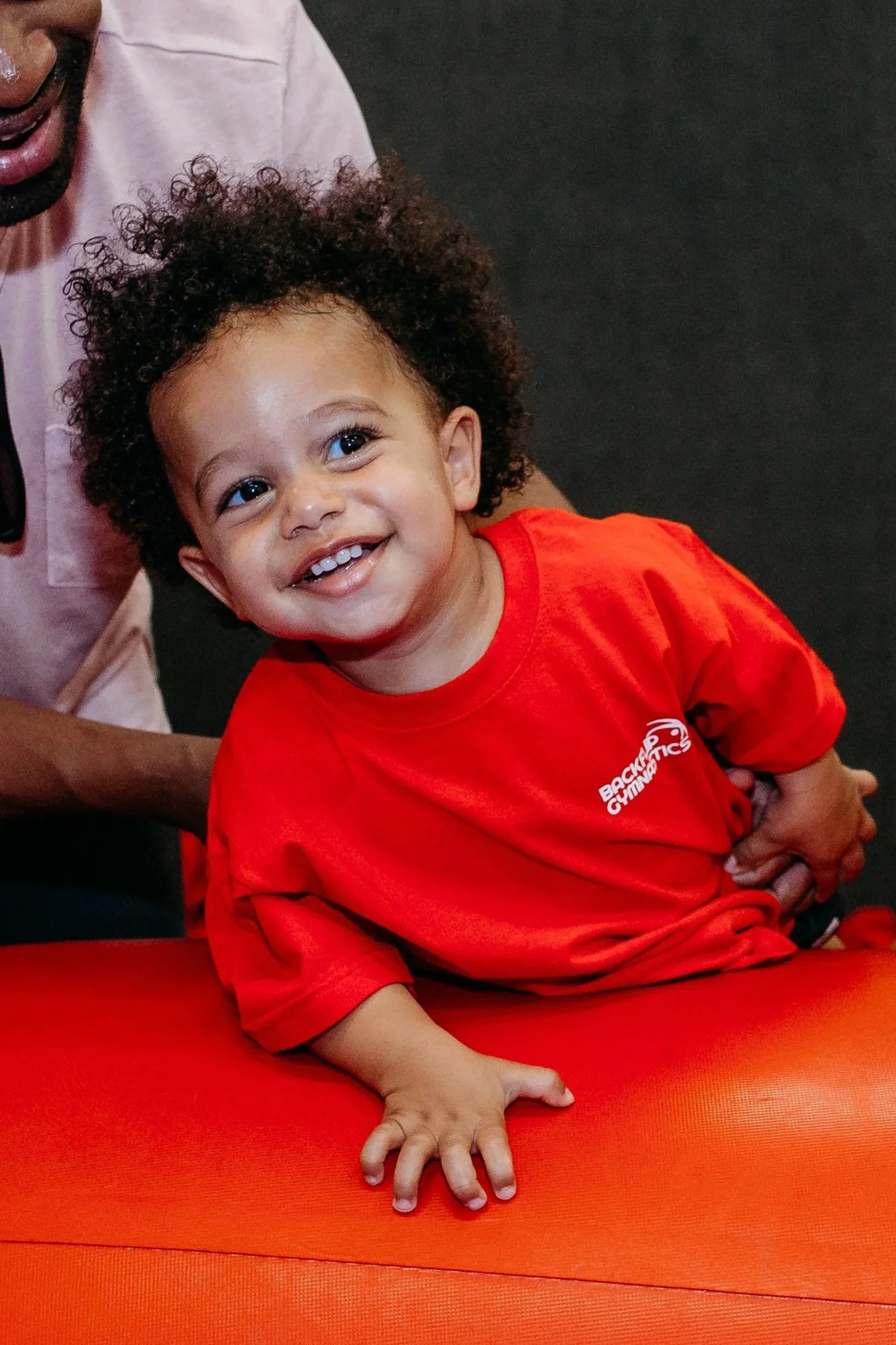 A young boy with curly hair smiling and leaning on a red exercise mat, wearing a red t-shirt with a small logo. An adult is partially visible holding him.