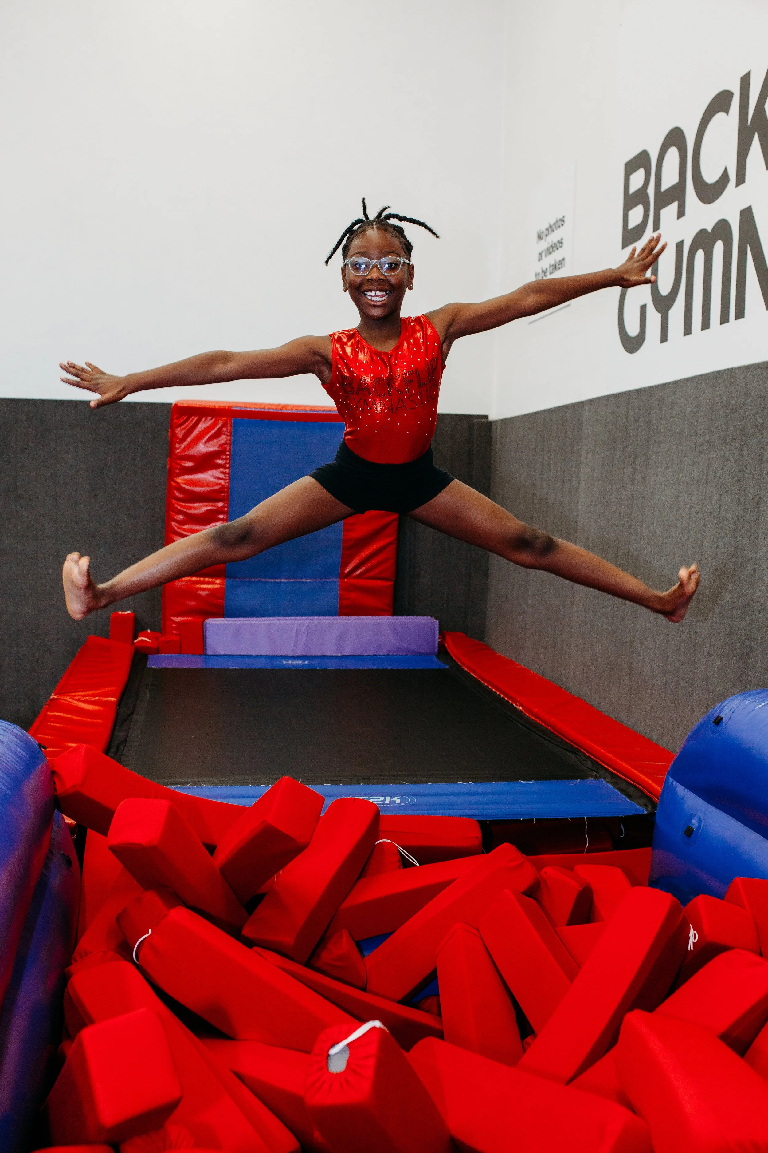 Young girl in a red gymnastics outfit jumping in mid-air on a trampoline at an indoor trampoline park.