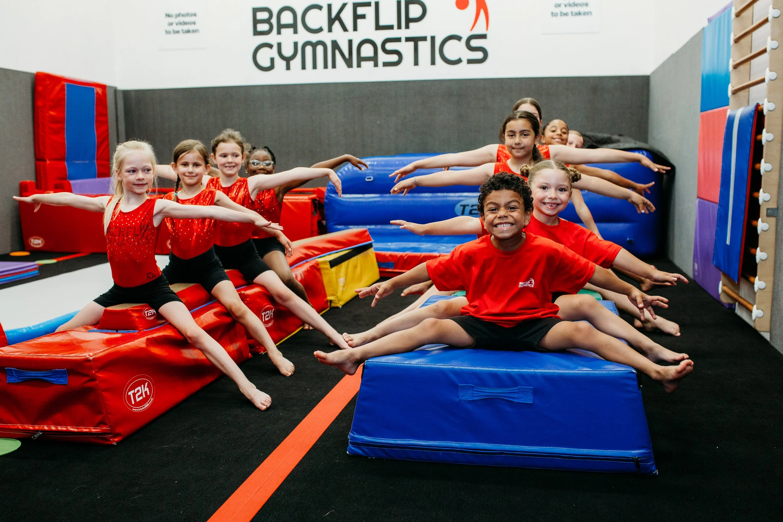 A group of young children with diverse skin tones wearing red and black gymnastics outfits, sitting in a split position on gym mats, with arms extended to the sides, smiling in a gymnastics gym.