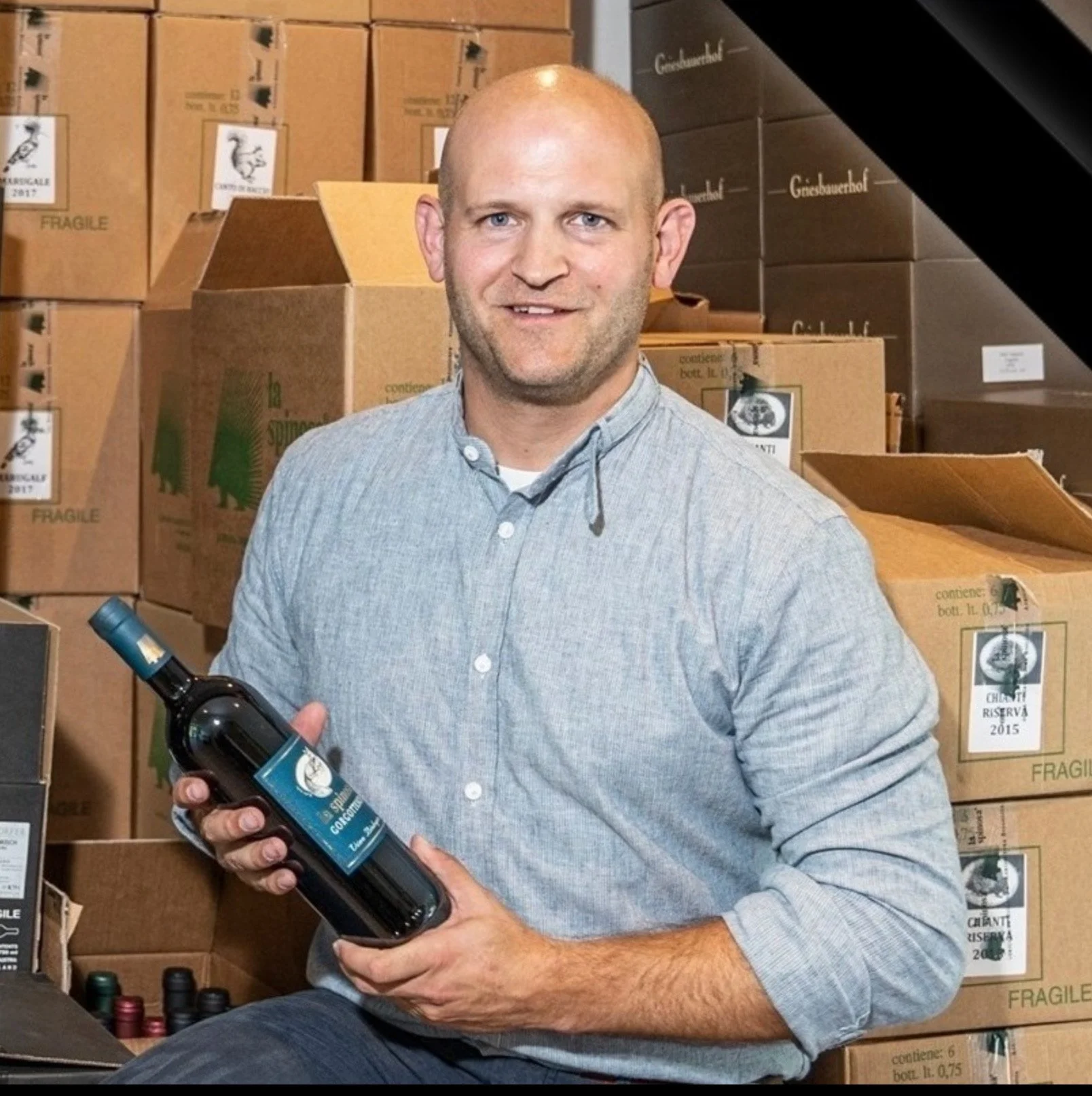 Ein Mann mit blauen Augen und Glatze hält eine Flasche Wein in den Händen, im Hintergrund sind Kartons und Weinflaschen zu sehen.