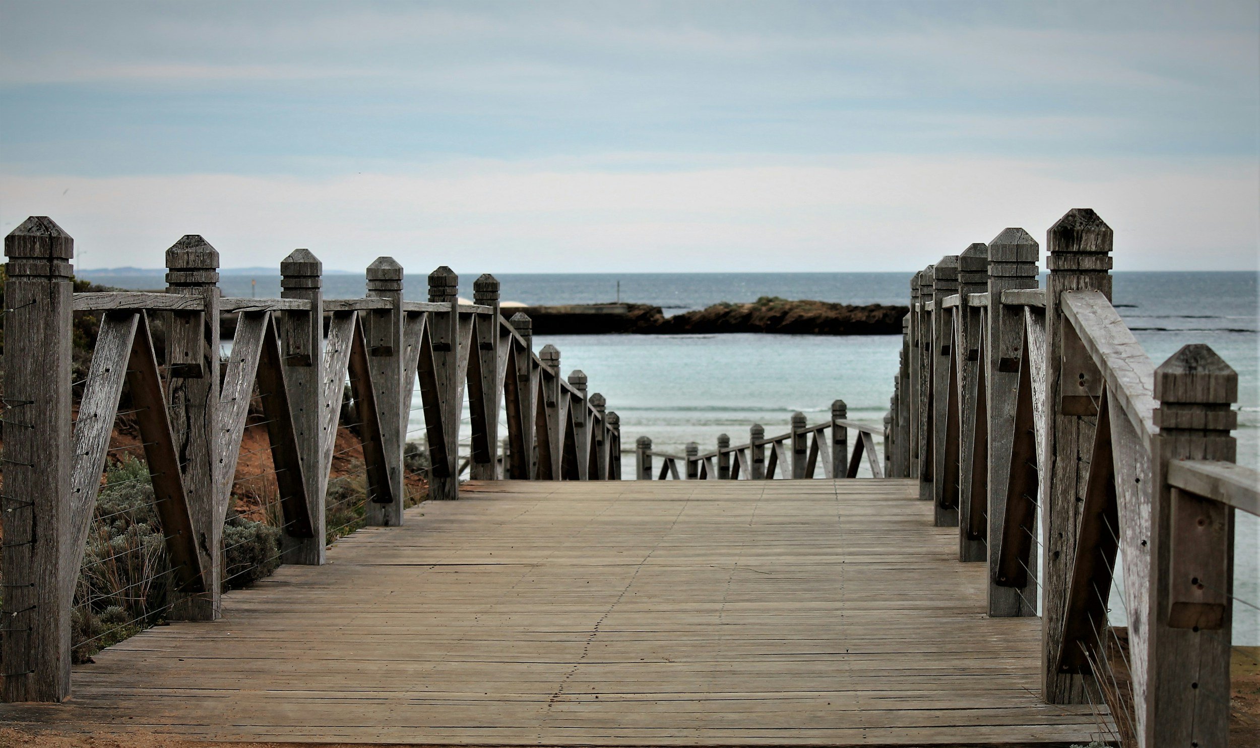 Wooden walkway leading to the ocean, with wooden railings on both sides and rocks in the distance.