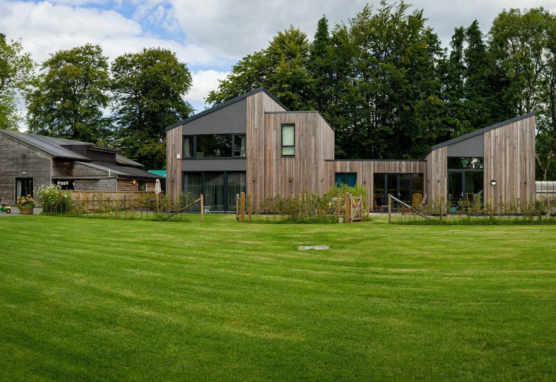 Modern house with wooden exterior and large glass windows, surrounded by a well-manicured green lawn and tall trees in the background.