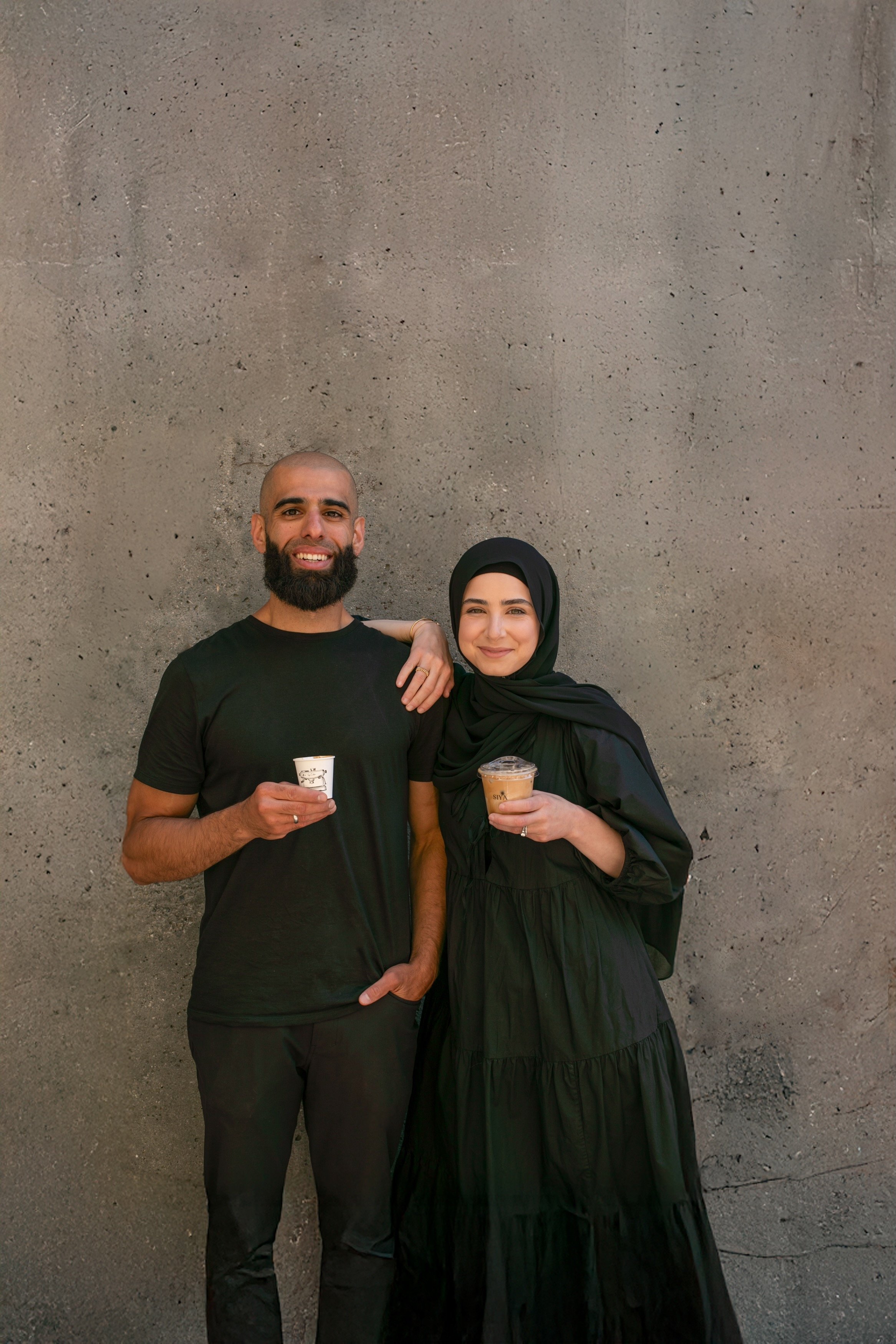 A man and woman standing against a concrete wall, both holding drinks and smiling at the camera.