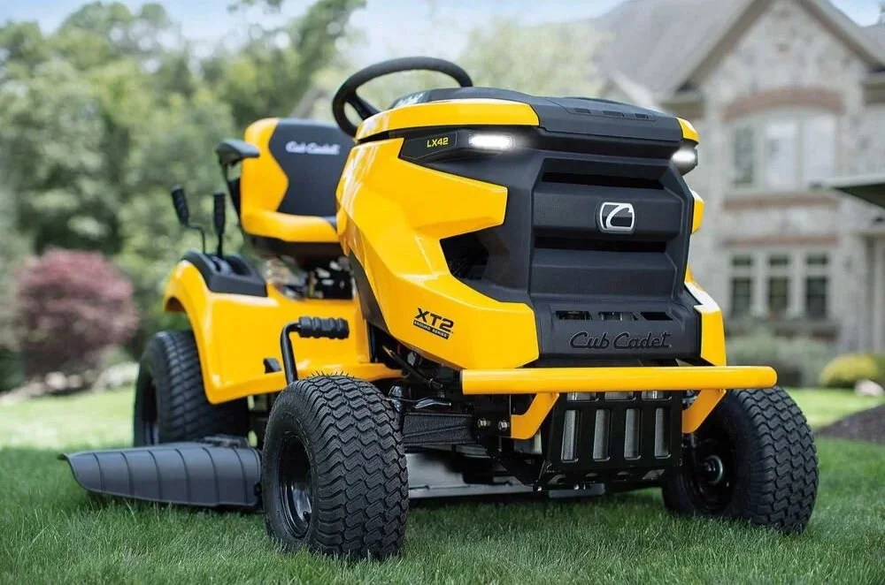 Yellow Cub Cadet XT2 riding lawn mower on a grassy yard with a house and trees in the background.