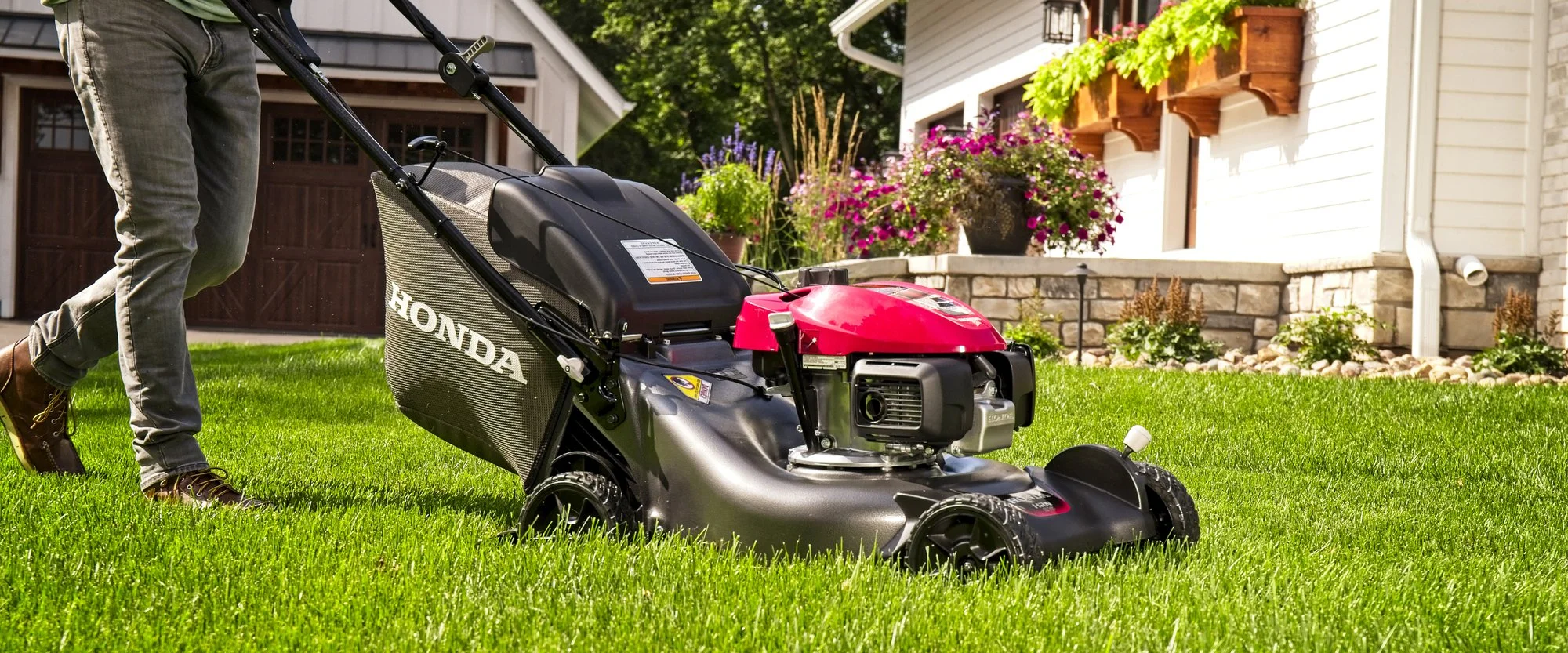 A person mowing a lush green lawn with a Honda push lawn mower in front of a house with flower beds and planters.