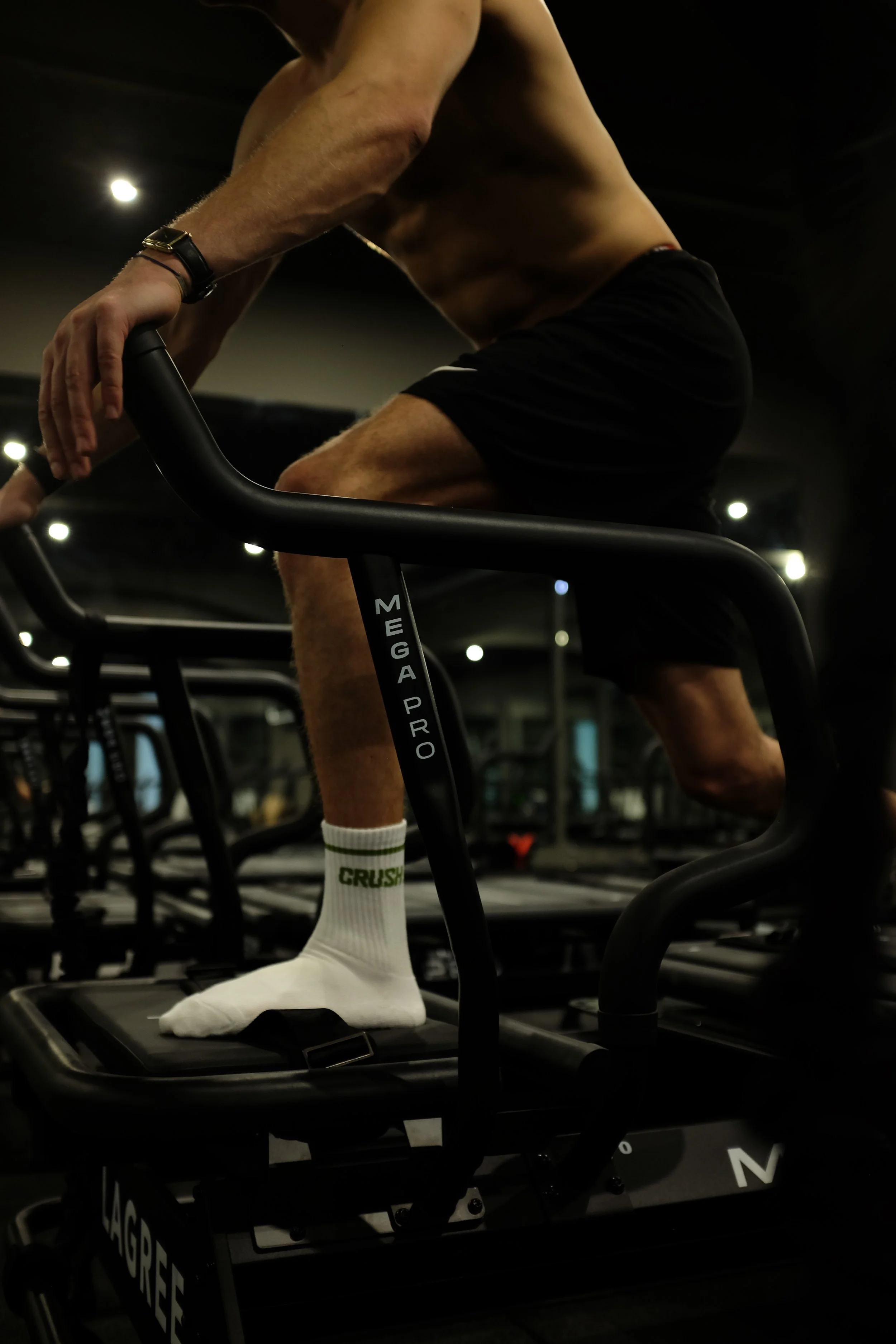 A shirtless man wearing black shorts and white socks with green text is exercising on a Mega Pro Lagree machine in a Lagree gym in vienna, holding onto a handrail.