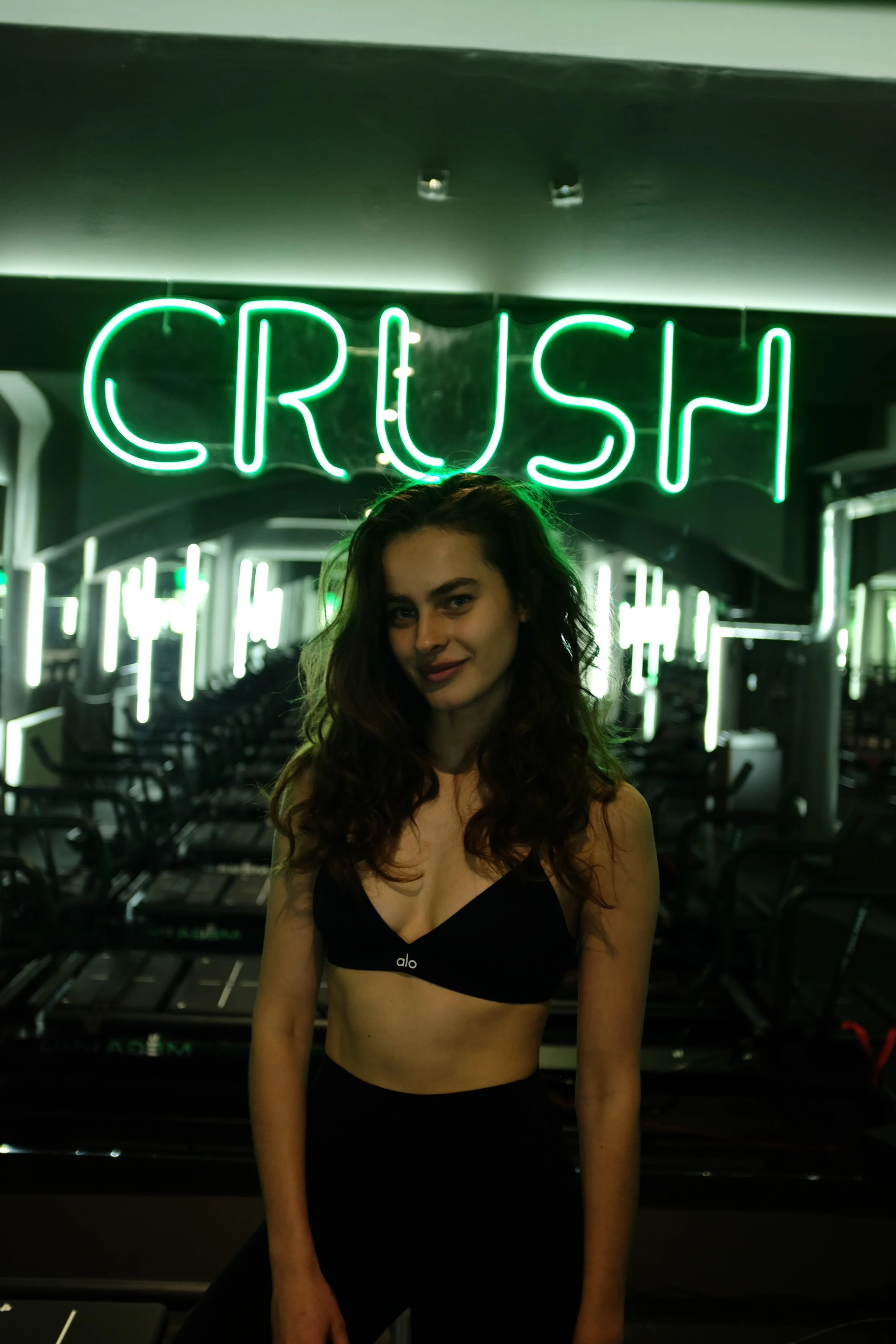 A young woman with long curly hair smiling in front of a neon green sign that reads 'CRUSH' inside a gym, with Lagree machines in the background.
