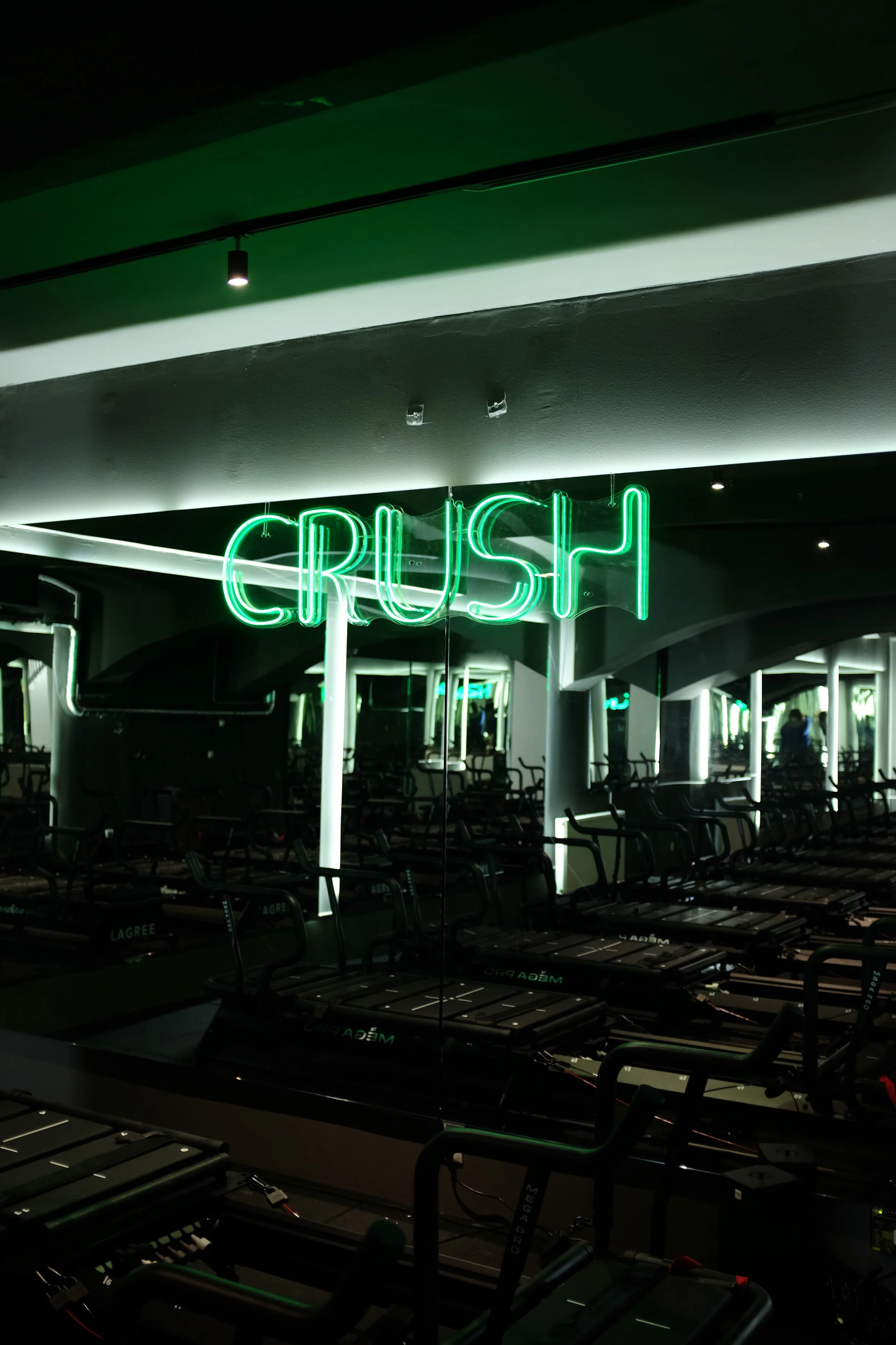 Neon green sign spelling 'CRUSH' in a gym with Lagree Megaformer and mirrors.