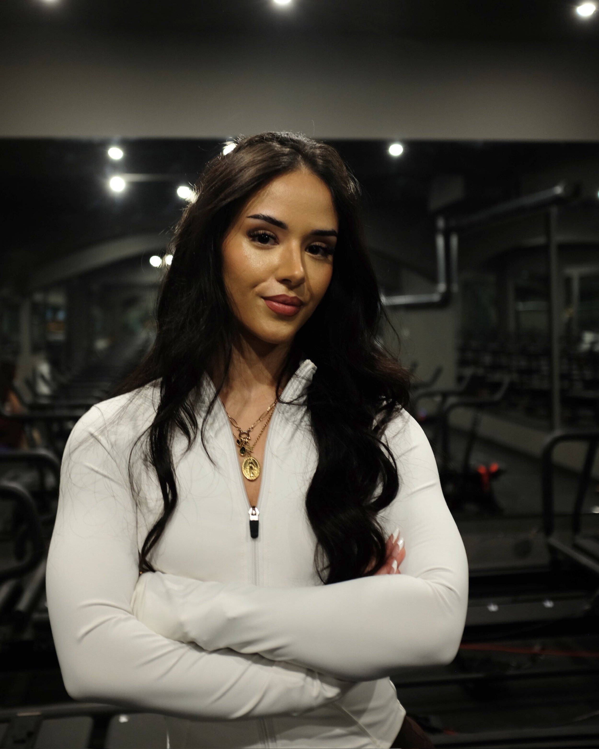 A young woman with long dark wavy hair, wearing a white zip-up athletic jacket and gold jewelry, standing with arms crossed in a gym with Lagree Machines in the background.