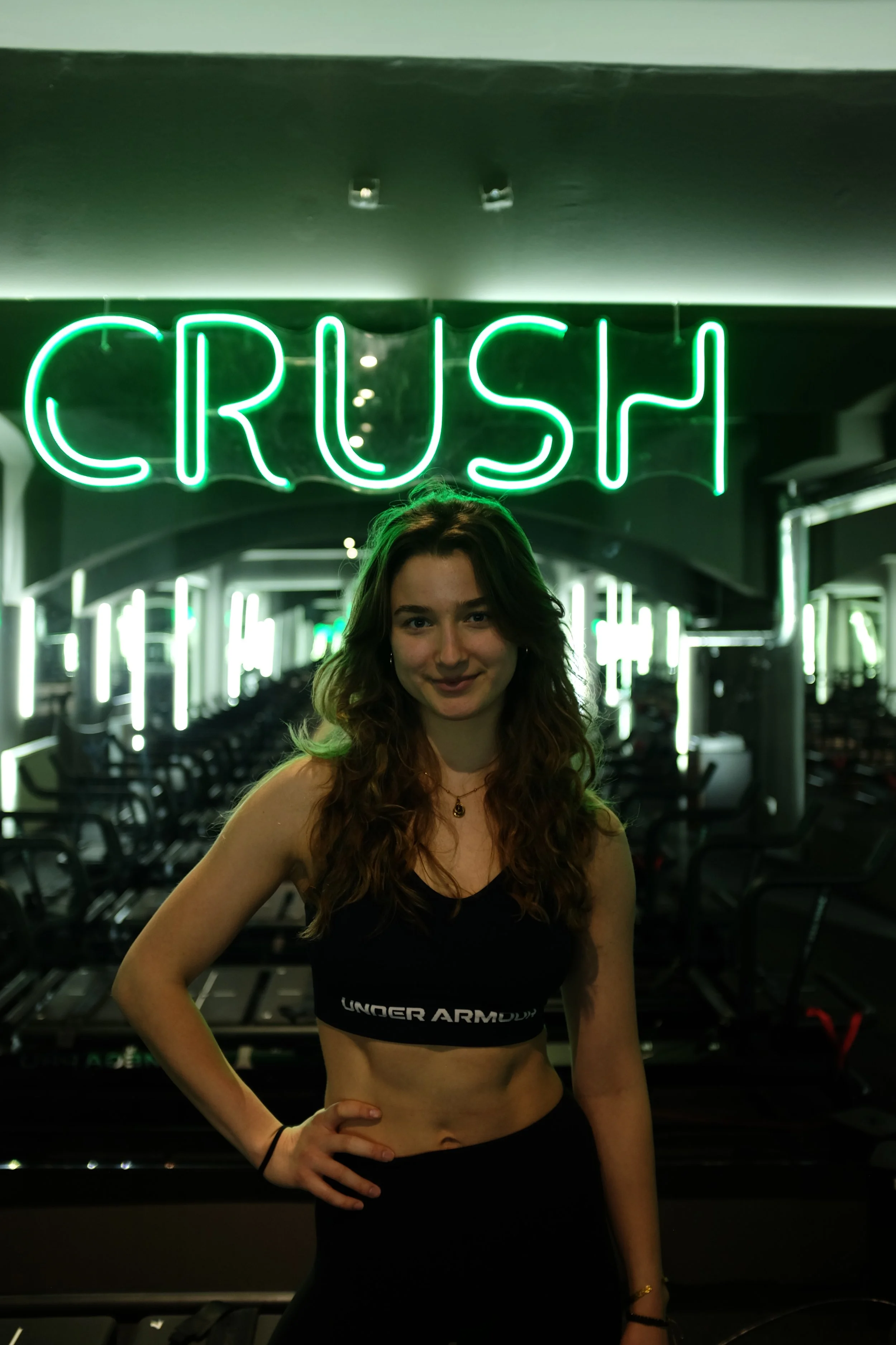 A young woman stands in front of a bright green neon sign that says 'CRUSH' inside a gym, wearing a black sports bra and black workout pants.