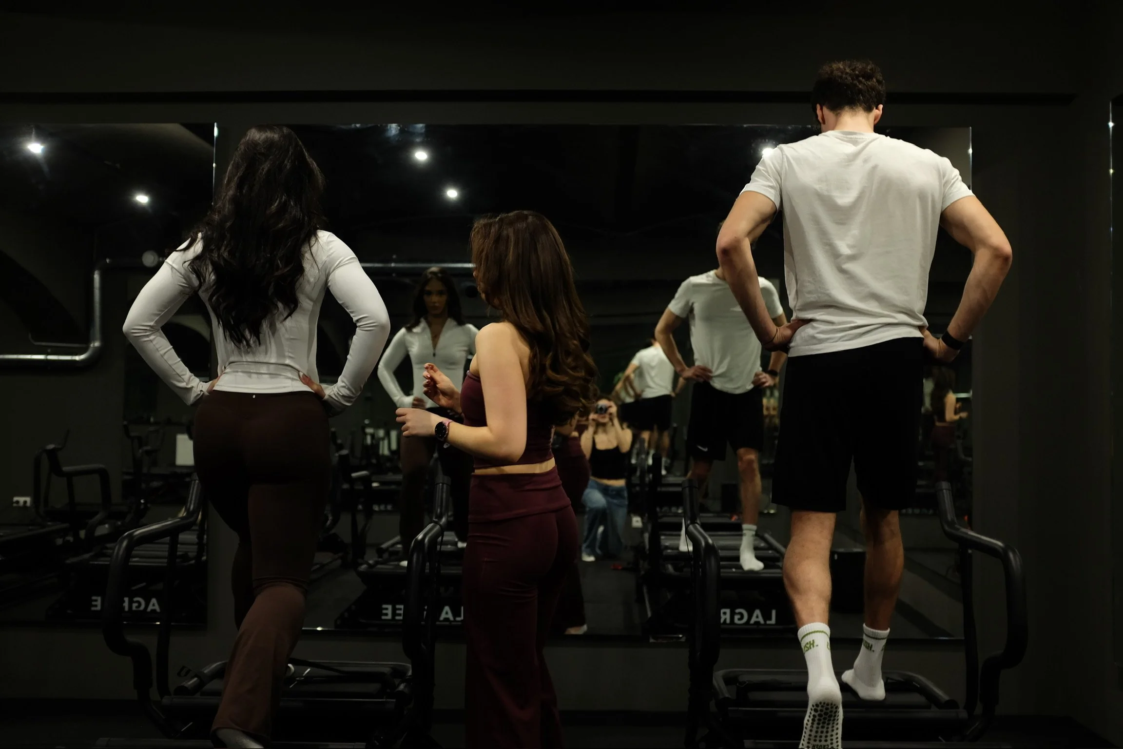 People at a gym, with one person looking at themselves in a mirror, others standing, exercising, or talking.
