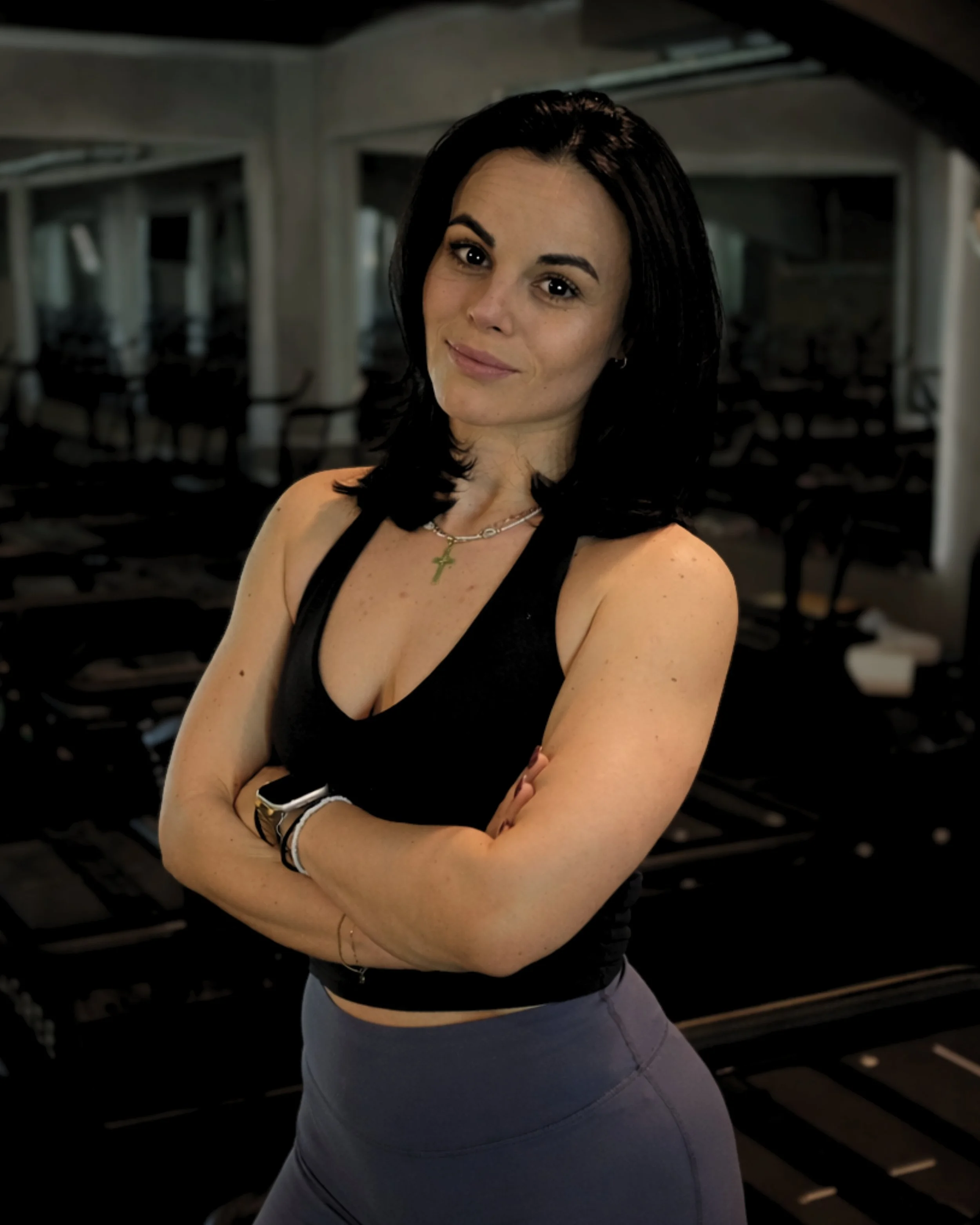 A woman with dark hair in a black tank top and gray leggings posing with arms crossed in a dimly lit indoor setting.