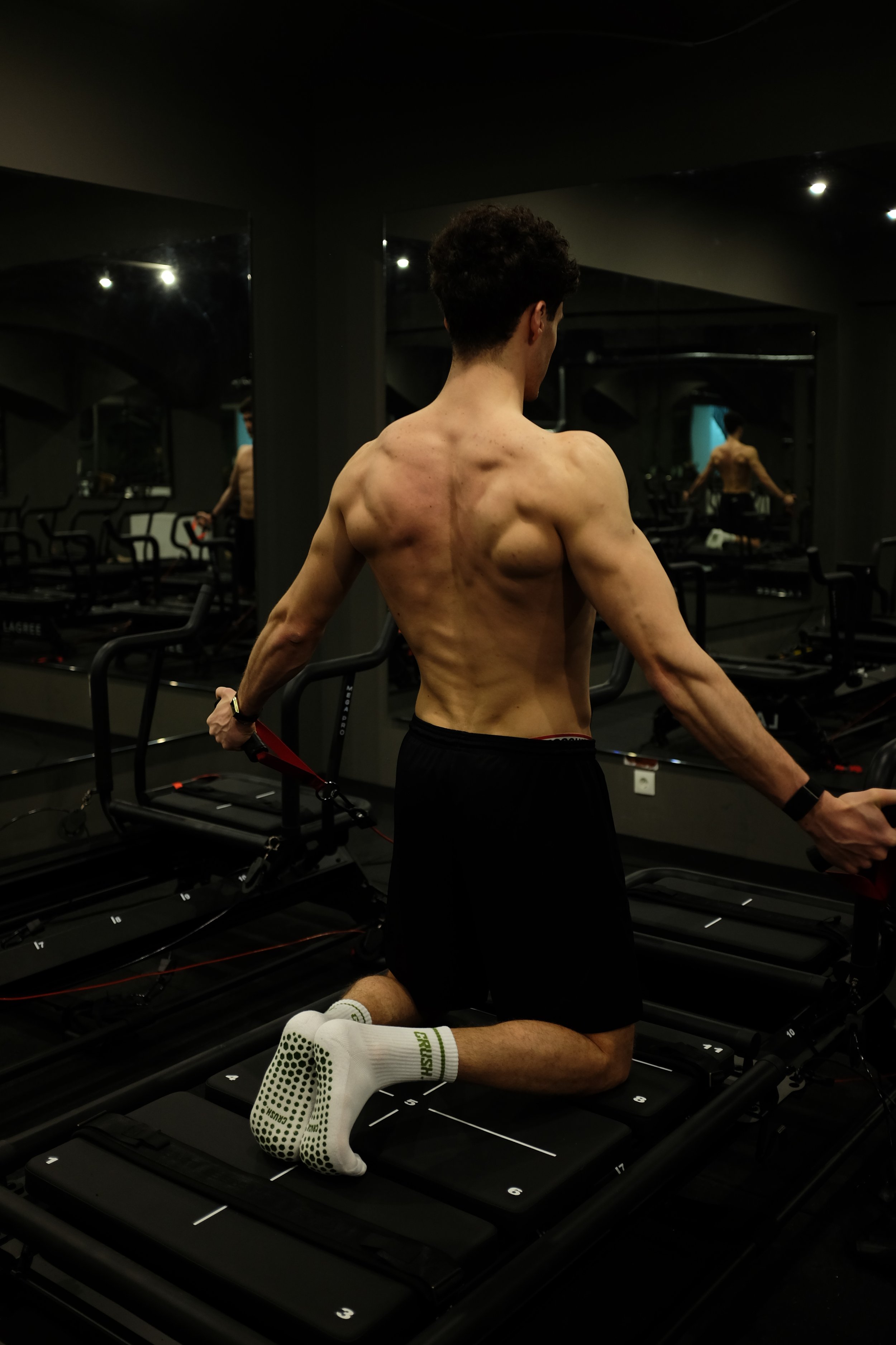 A shirtless man exercising on a treadmill in a gym, with mirrors reflecting his back muscles and fitness. He is kneeling on the Lagree Machine with his hands holding the handles.