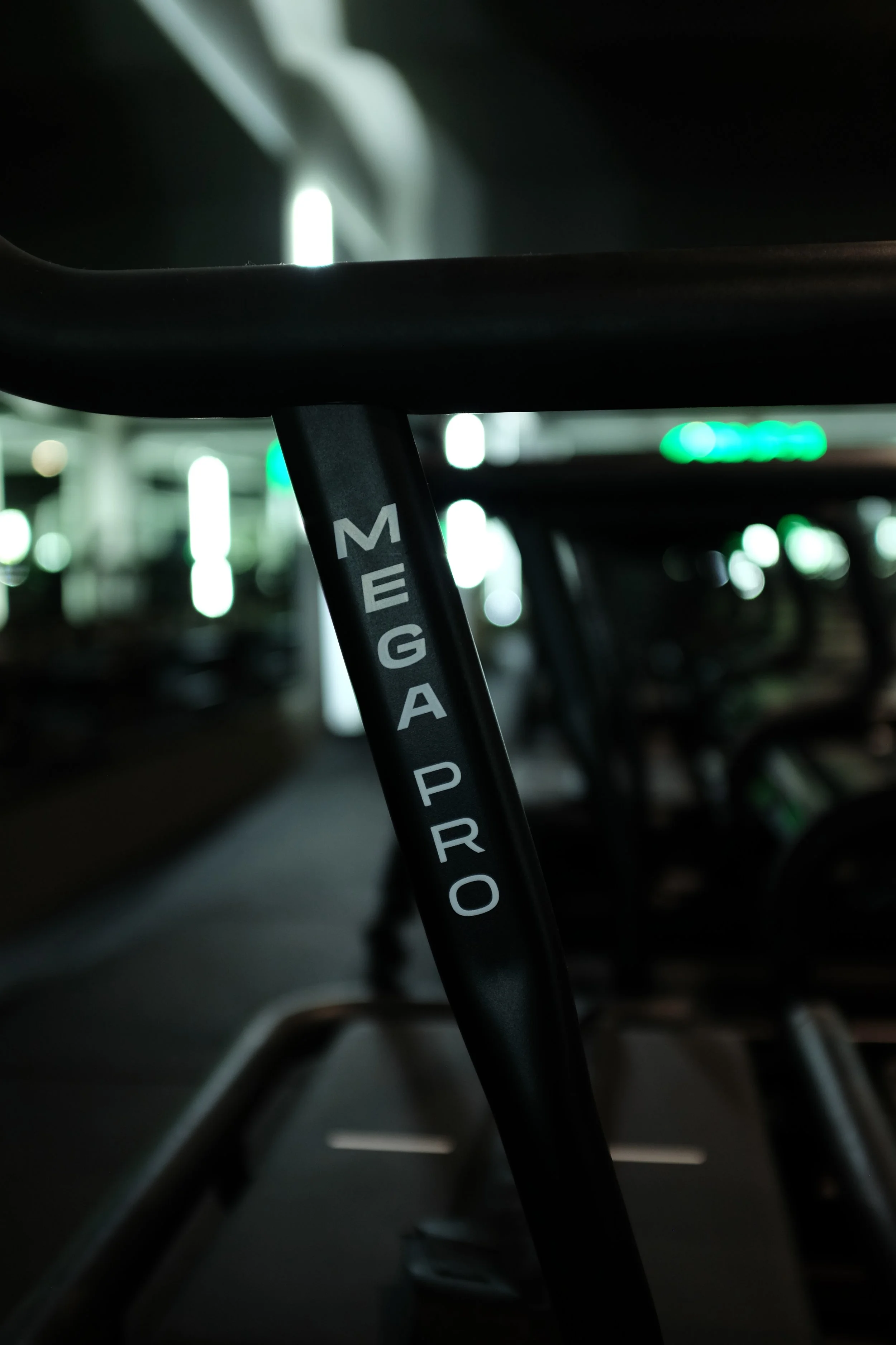 A black Lagree Machine at a lagree fitness gym in vienna with a close-up of the machine handles that says 'MEGA PRO' in white lettering, with the background showing a blurred lagree fitness gym interior with bright lights.