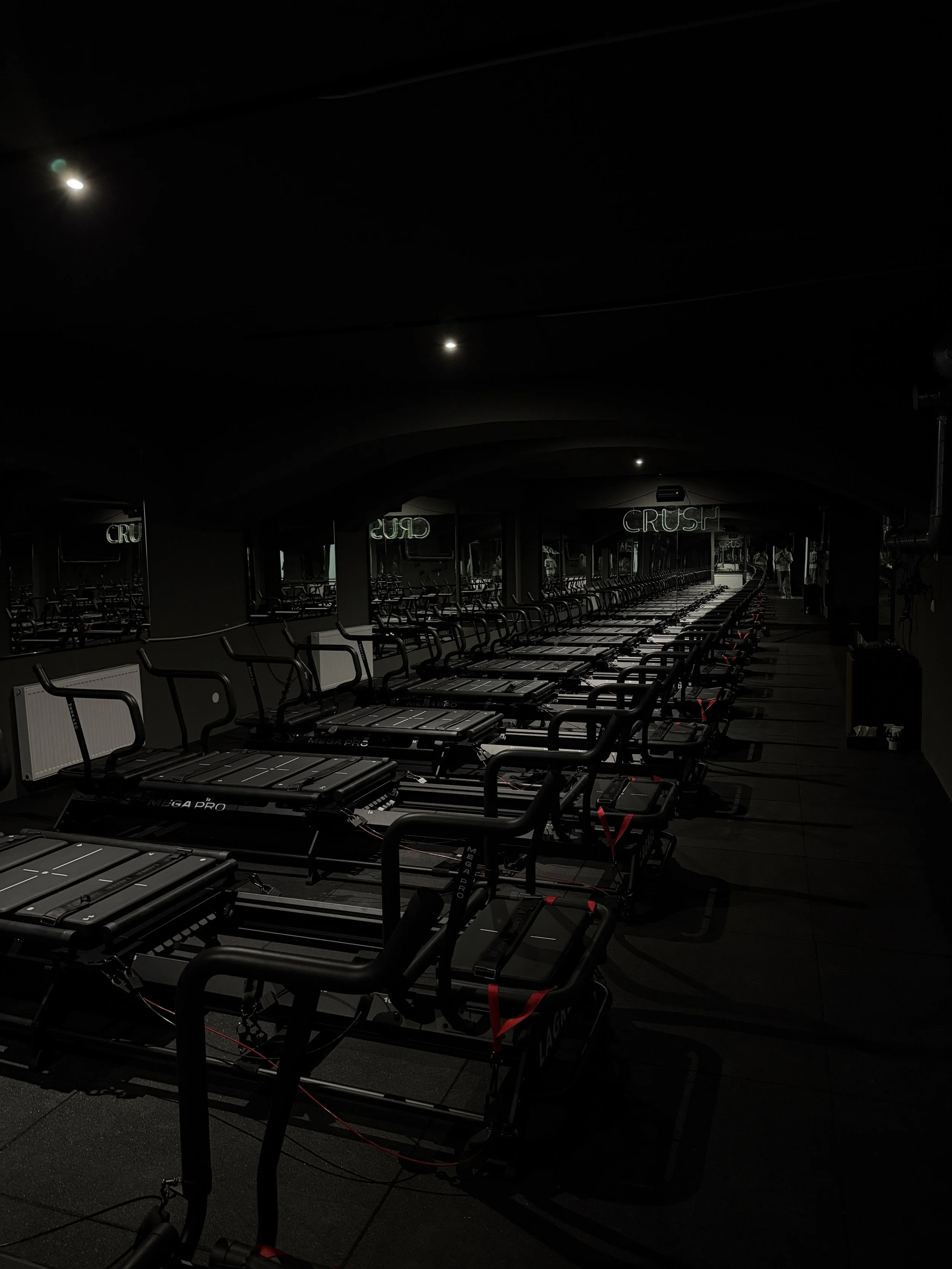 Leerer Raum mit mehreren Laufbändern im Dunkeln, Spiegel an der Wand, Neon-Schriftzug 'CRUSH' an der Decke, Reflektionen der Laufbänder in den Spiegeln.