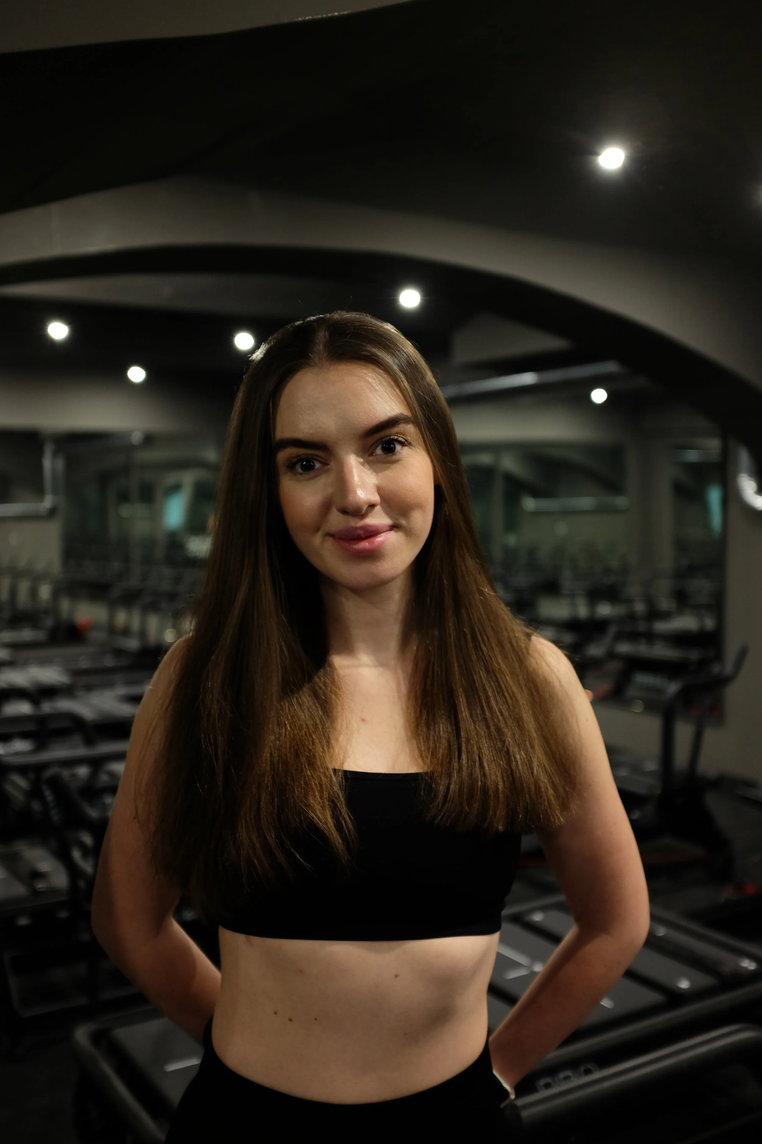 A young woman with long brown hair, wearing a black sports top, standing in a dimly lit gym, with Lagree machines in the background.
