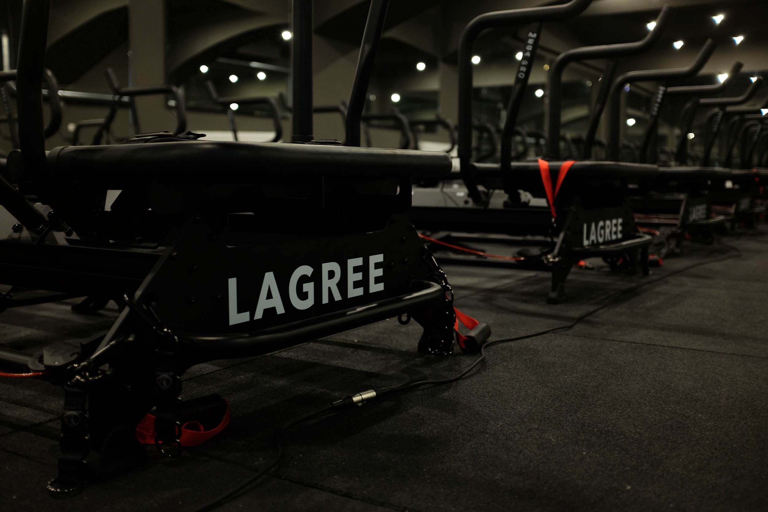 Row of black Lagree Megaformer machines labeled 'LAGREE' in a dimly lit indoor rowing facility.