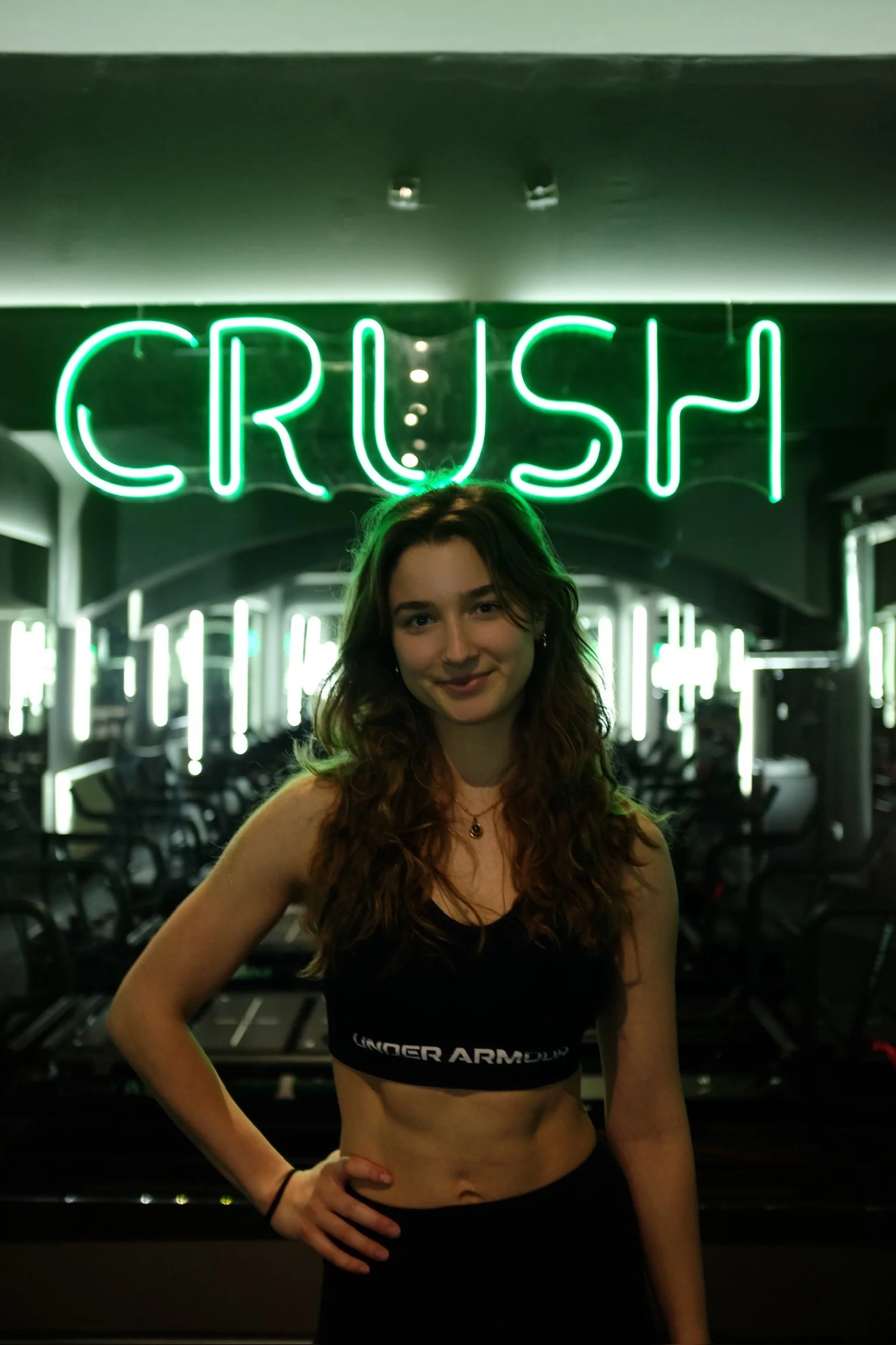 Eine junge Frau mit braunen, lockigen Haaren posiert vor einer Neonwerbetafel mit der Aufschrift 'CRUSH'. Sie trägt ein schwarzes Unterwäsche-Oberteil mit dem Markenname 'Under Armour' und eine schwarze Hose, im Hintergrund sind Tische und Stühle sichtbar.
