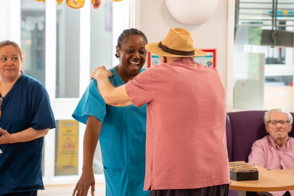 A woman in blue scrubs receives a warm hug from an elderly man wearing a pink shirt and a straw hat, while another woman in blue scrubs looks on nearby in a brightly lit room.