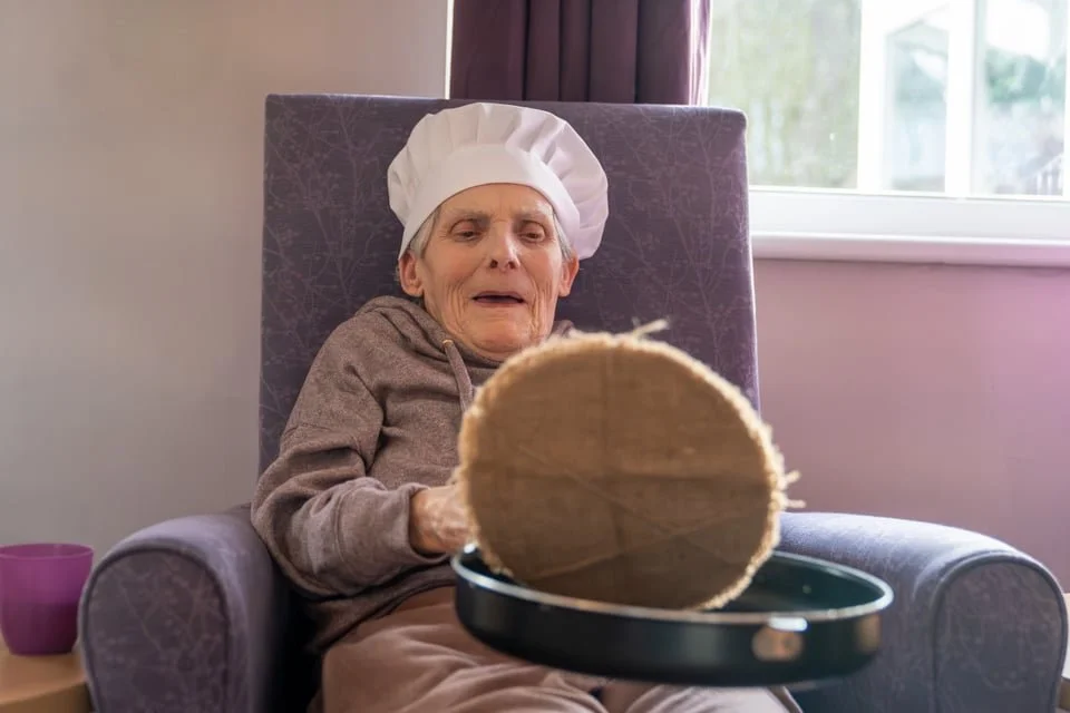 An elderly woman sitting in a purple armchair wearing a chef hat, looking at a cheesecake with a surprised expression.