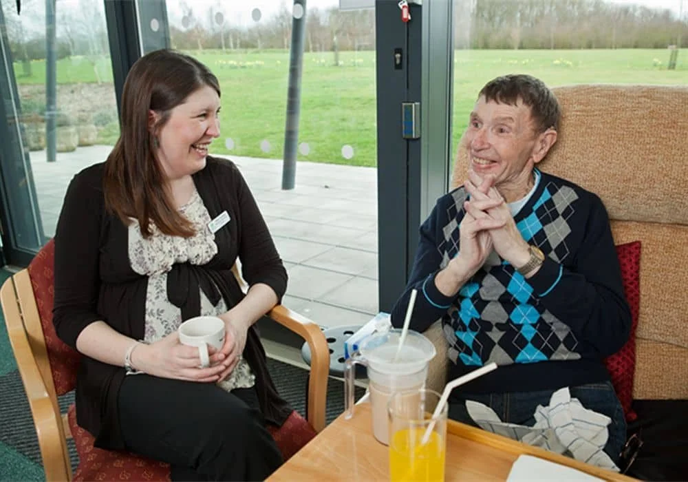 A woman and an elderly man sitting inside a cafe, smiling and enjoying conversation. The woman is holding a mug, and there are drinks and a straw on the table.